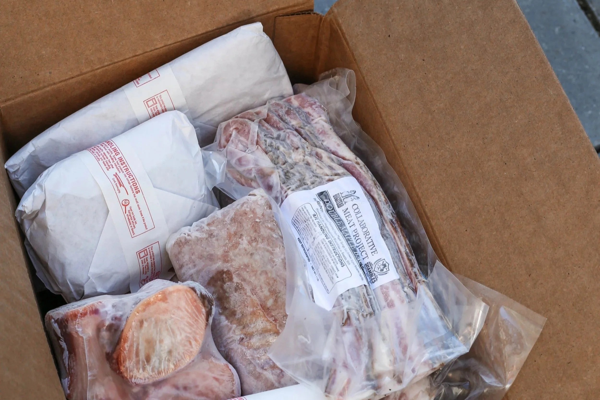 An open cardboard box filled with various cuts of meat, including vacuum-sealed ribs and packages wrapped in white butcher paper.