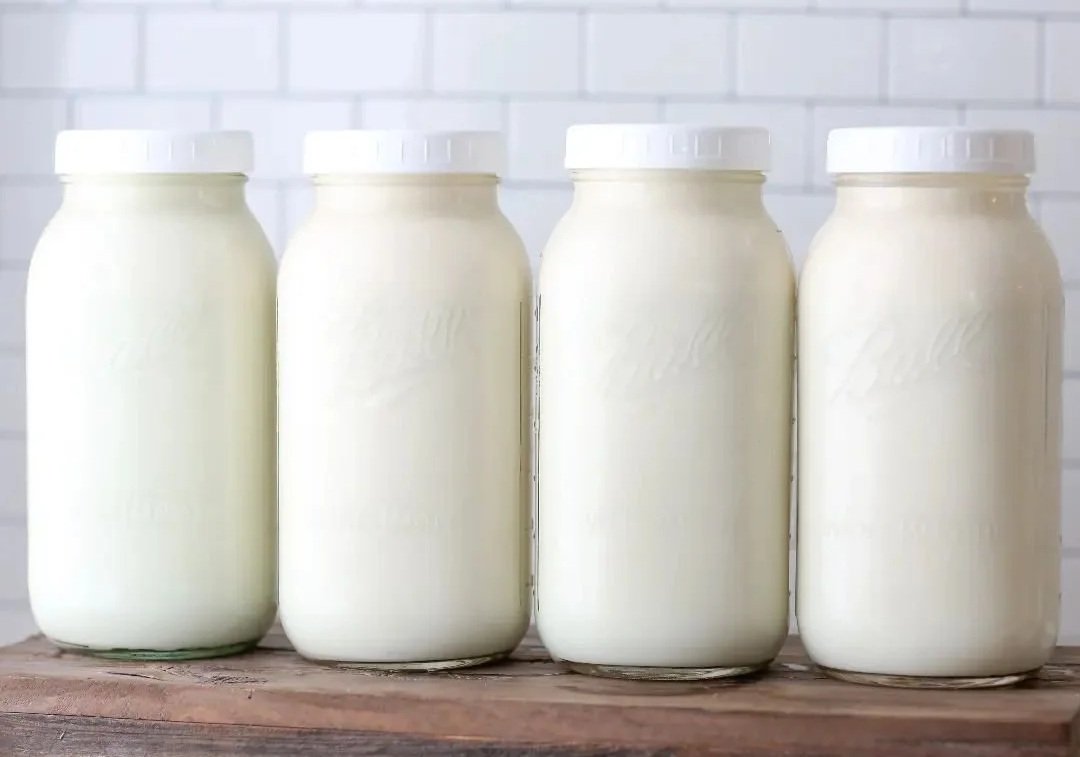 Four glass jars filled with fresh milk lined up on a wooden surface against a white tile background.