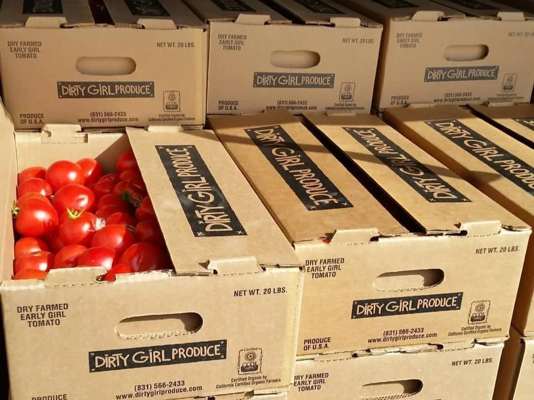 Stacked cardboard produce boxes labeled "Dirty Girl Produce," with one open box displaying bright red tomatoes.