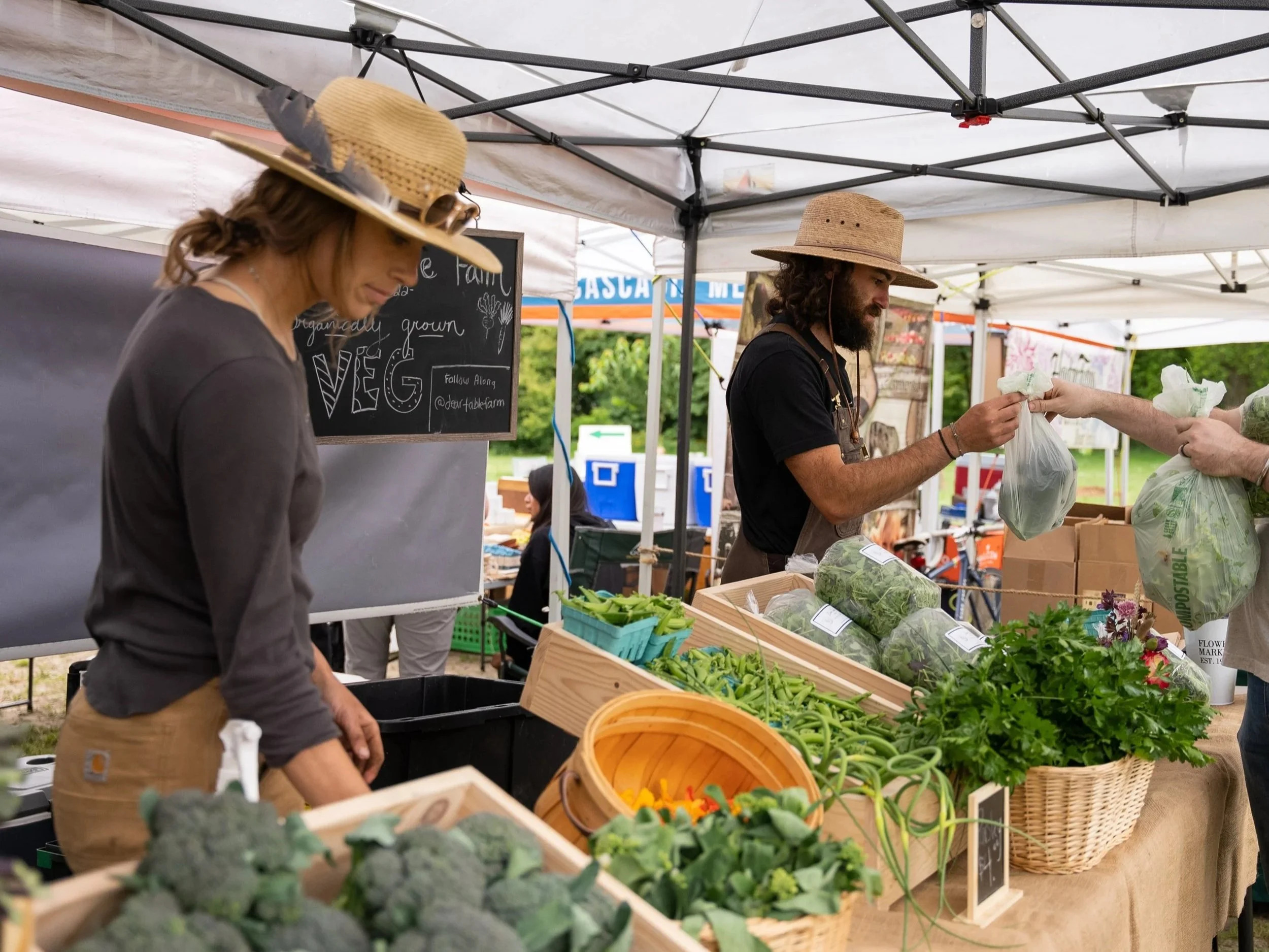 Farmers market vendors wearing straw hats selling fresh produce under a canopy tent.