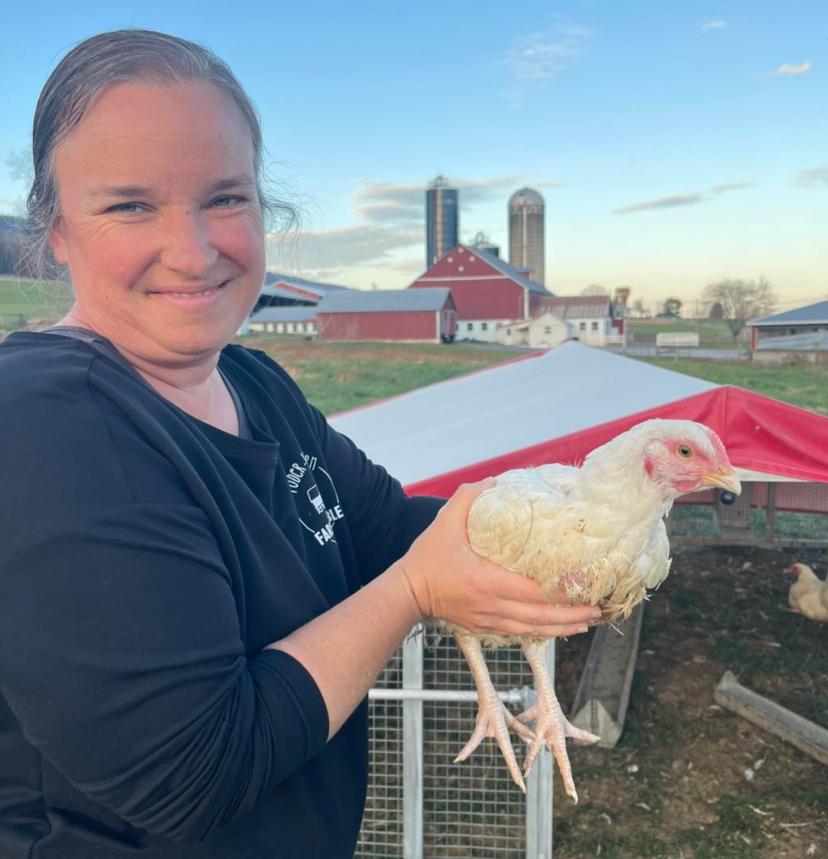 A smiling woman holding a white chicken outdoors on a farm with barns and silos in the background.
