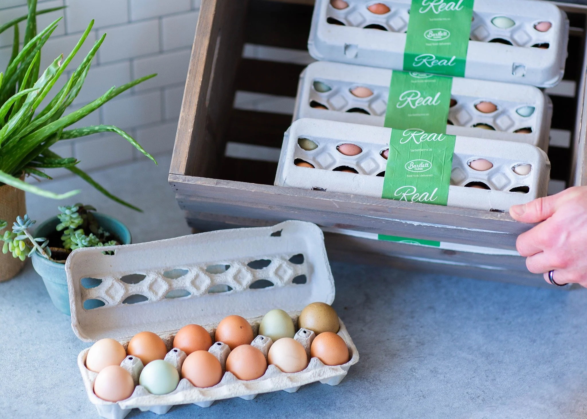An open carton of multi-colored farm eggs next to a wooden crate stacked with more egg cartons.