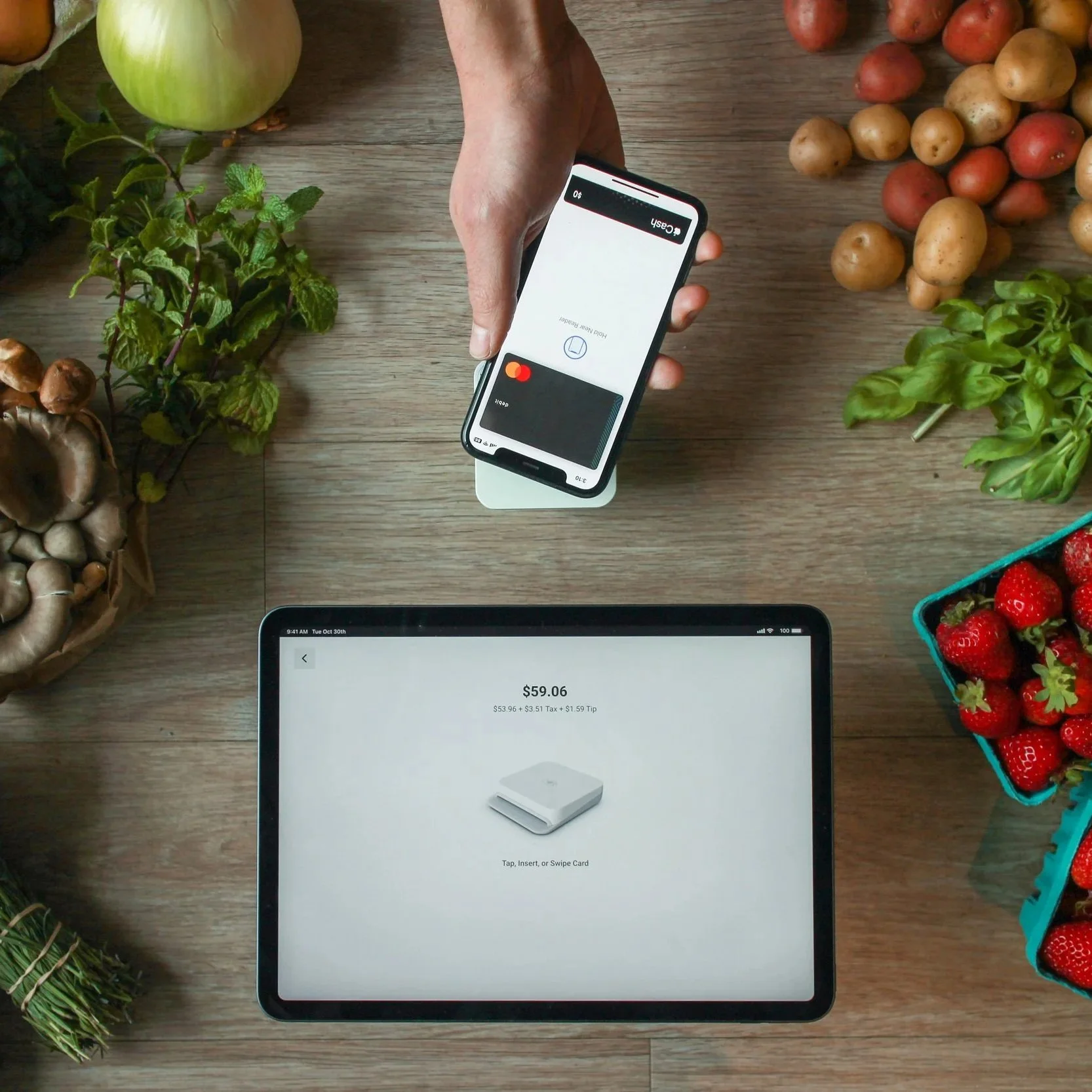 An overhead view of a digital payment being made at a produce stand. A person holds a smartphone over a white contactless card reader to pay via Apple Pay.
