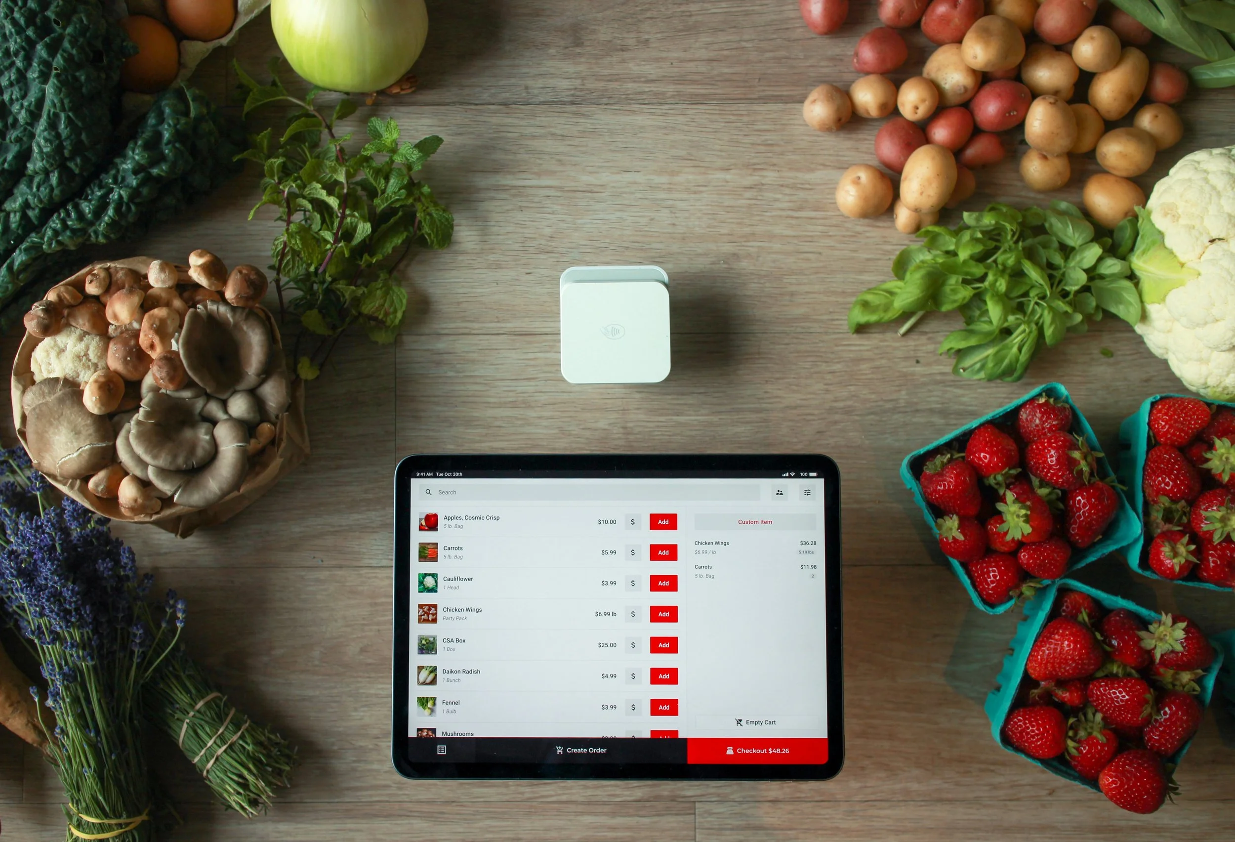 A top-down view of a digital tablet checkout screen and card reader surrounded by fresh produce, including strawberries, mushrooms, potatoes, and greens.