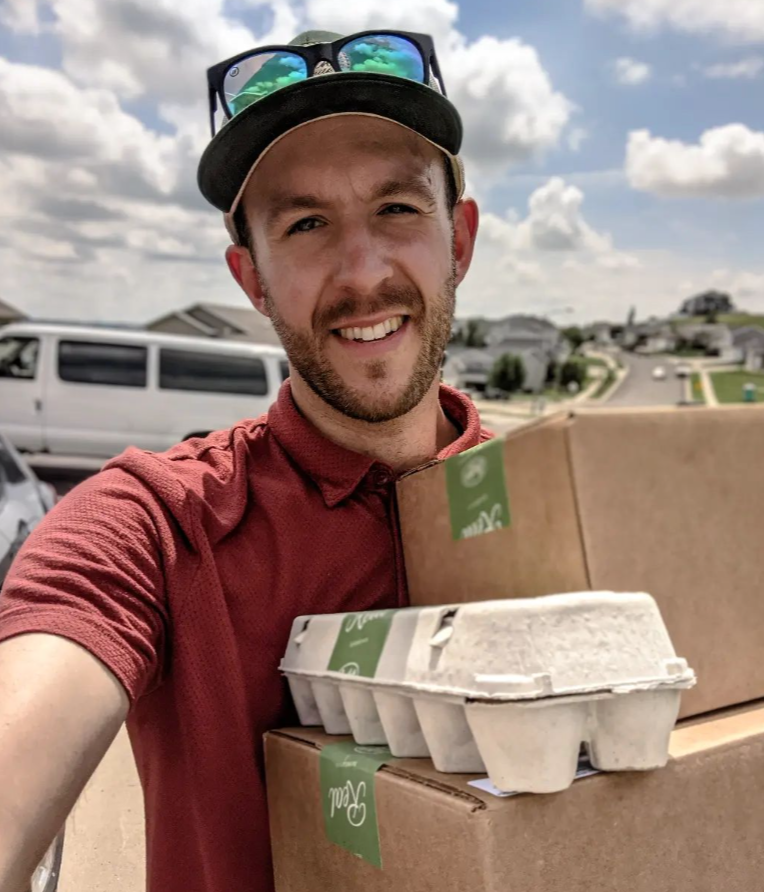 A smiling man holding cardboard delivery boxes and an egg carton outdoors.