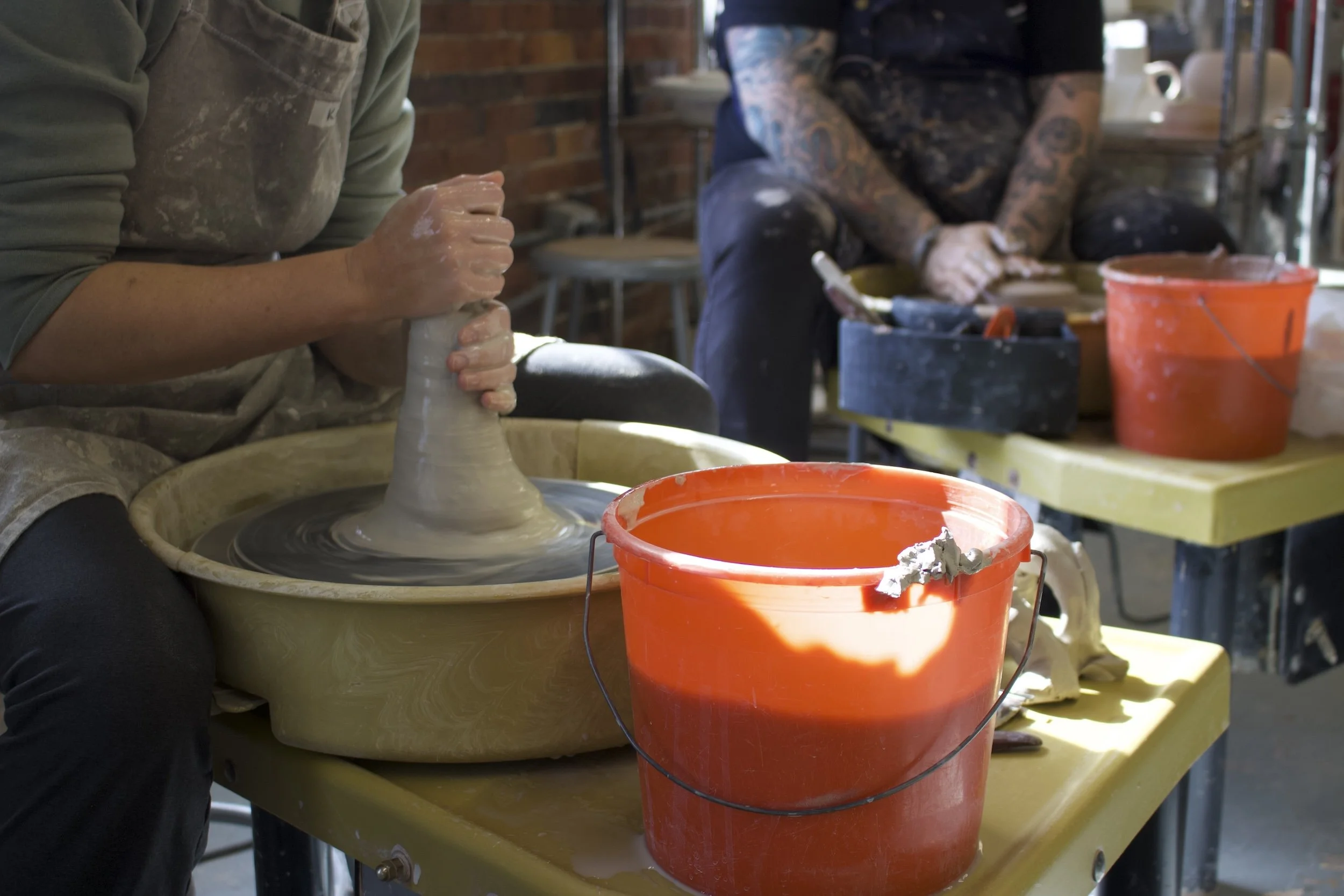 Person shaping clay on a pottery wheel with an orange bucket nearby, and another person sitting in the background with tattoos.