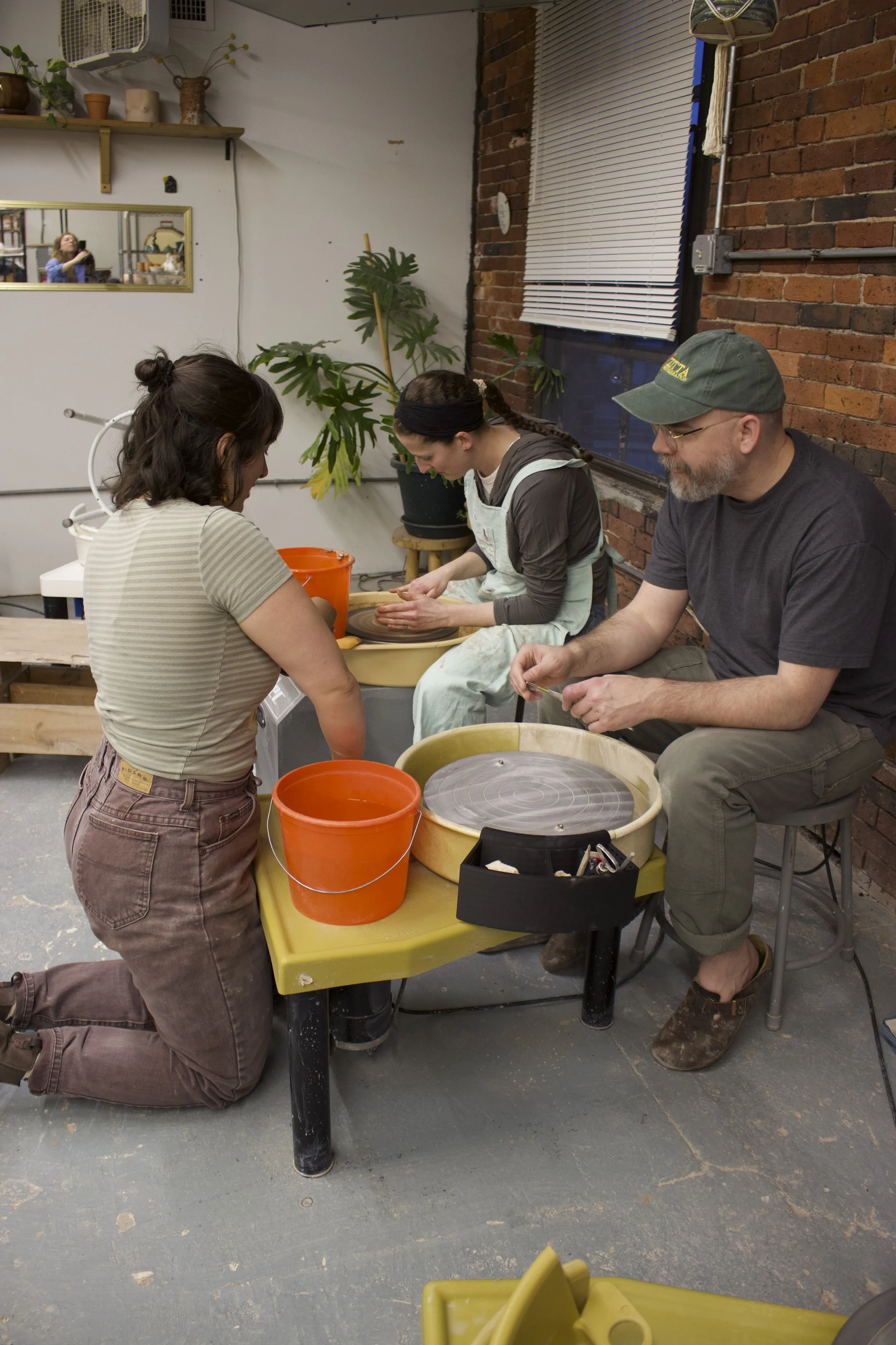People at pottery wheel creating clay pots in a studio with brick wall and plant in background.
