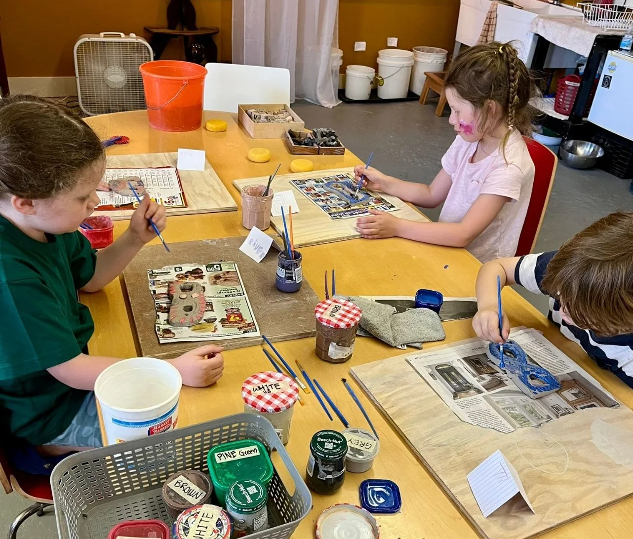 Three children sitting at a table painting and decorating ceramic ornaments, with various art supplies and jars of paint on the table.