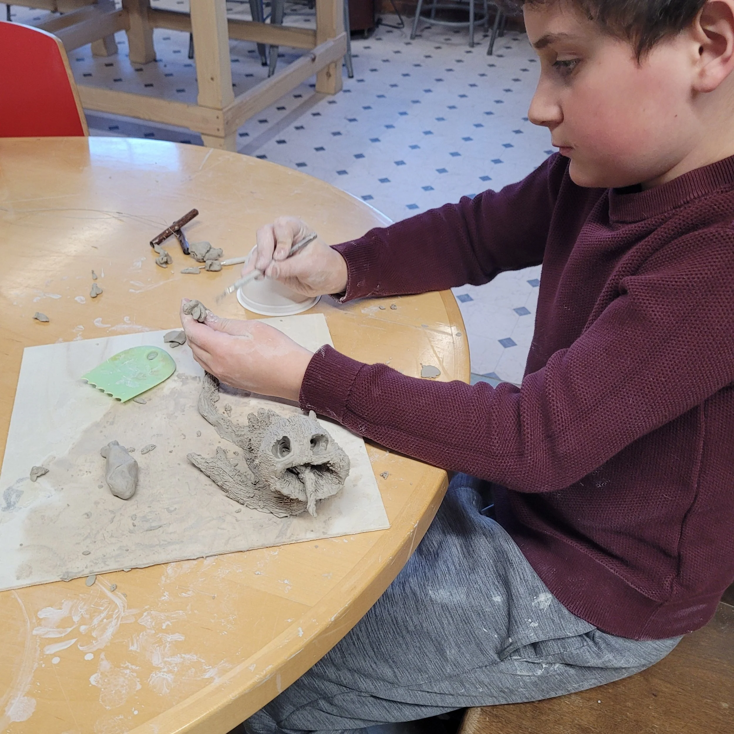 A person sculpting a lion's head out of clay at a round wooden table.