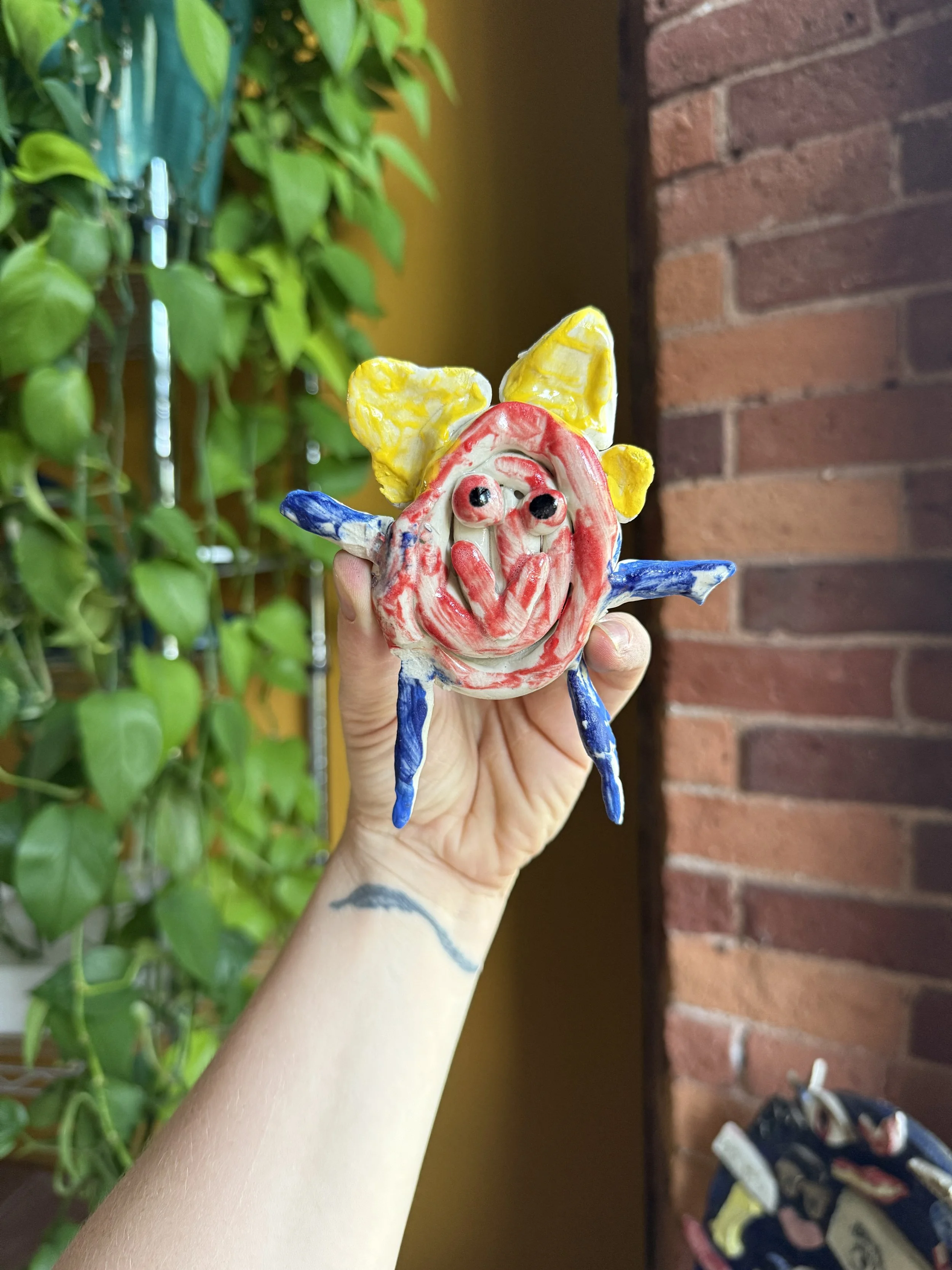 Colorful ceramic clown figurine being held up against a background of green plants and a red brick wall.