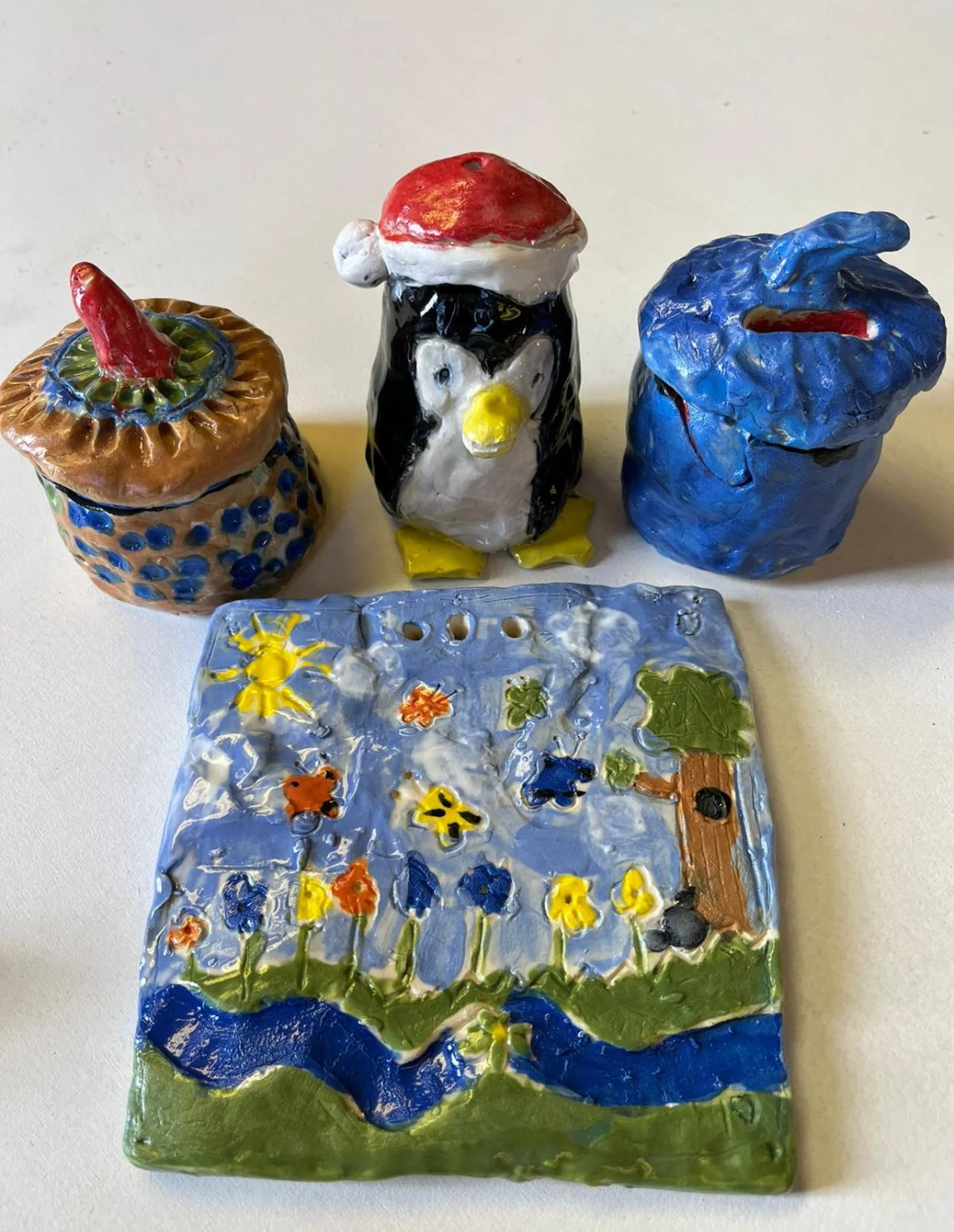 Three colorful ceramic figurines—a jar with a red chili on top, a penguin wearing a Santa hat, and an abstract blue sculpture—are arranged on a white surface, with a rectangular ceramic tile depicting a garden scene with flowers, a tree, and a blue river in front of them.