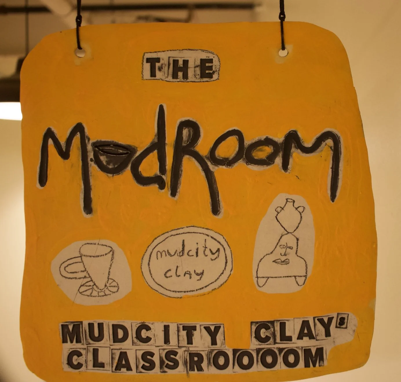 Sign reading "The Mudroom" with hand-drawn illustrations of a coffee cup, a spirit level, and a woman with a vase on her head, made from cut-out letters on a yellow background.