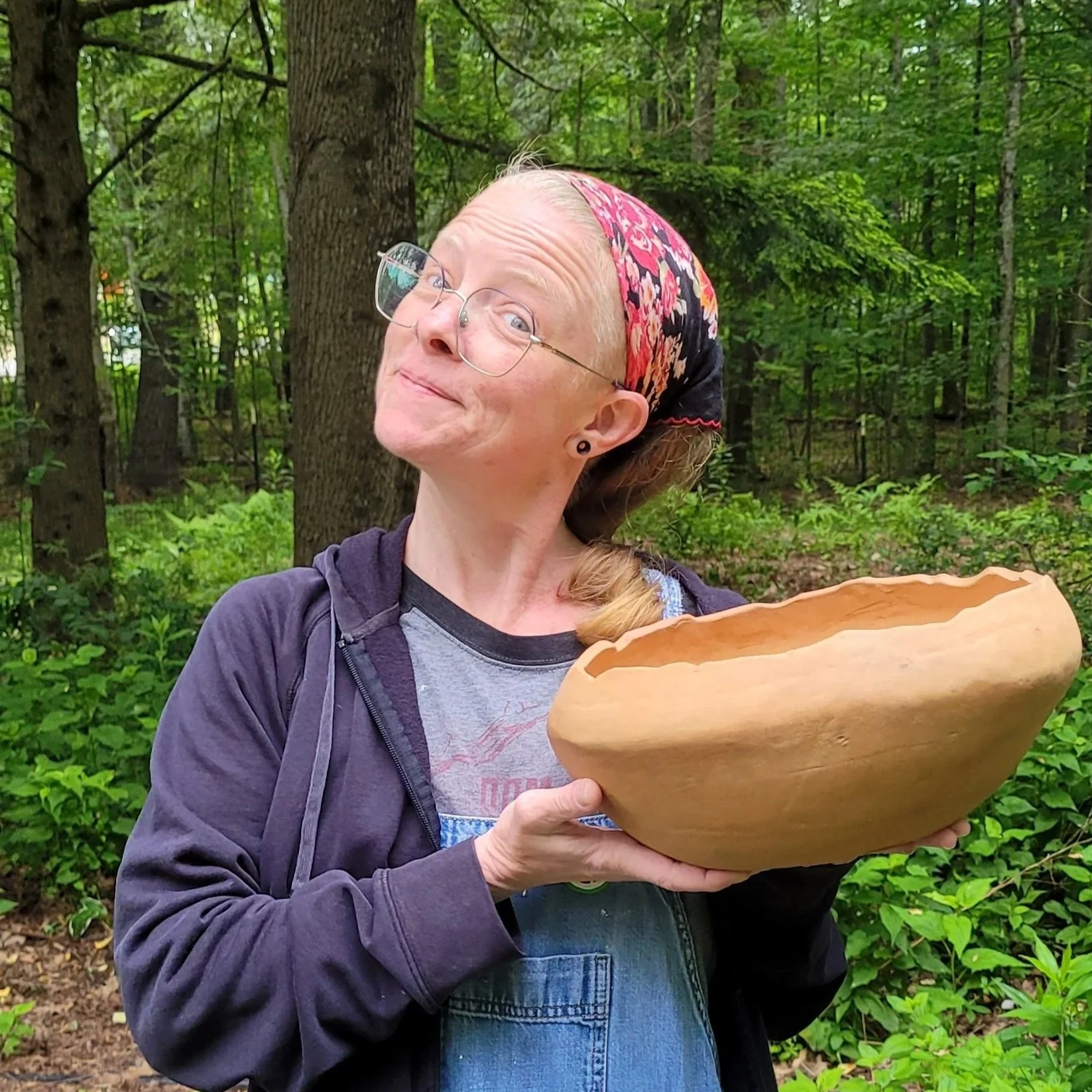 A woman with glasses, a colorful headscarf, and a dark jacket holding a large wooden bowl in a green forest.