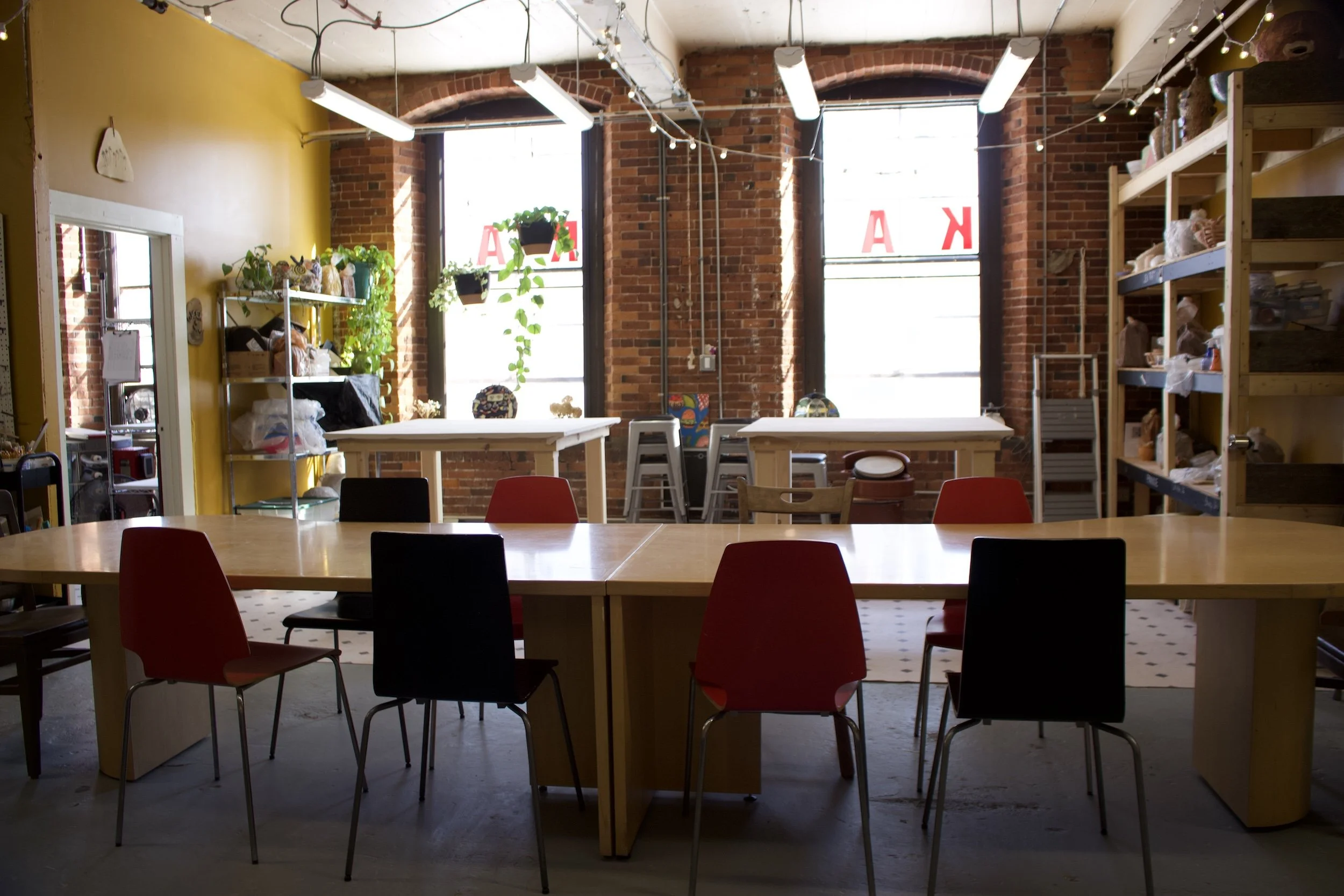A bright indoor space with a large wooden table surrounded by black and red chairs, brick walls, large windows with sunlight, shelves with plants and supplies, and string lights hanging from the ceiling.