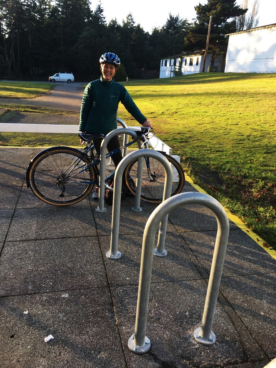 A woman standing next to her bicycle at a bike rack on a paved sidewalk, wearing a helmet and a teal jacket, with a grassy hill and white building in the background during sunset.