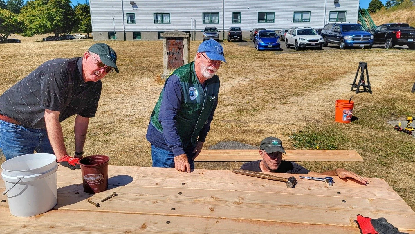 Three men are working on a wooden project outdoors on a sunny day. One man is inside the wood, holding a tool, while the other two are leaning over and observing. There are tools and supplies on the wood and in the background, cars are parked near a building.
