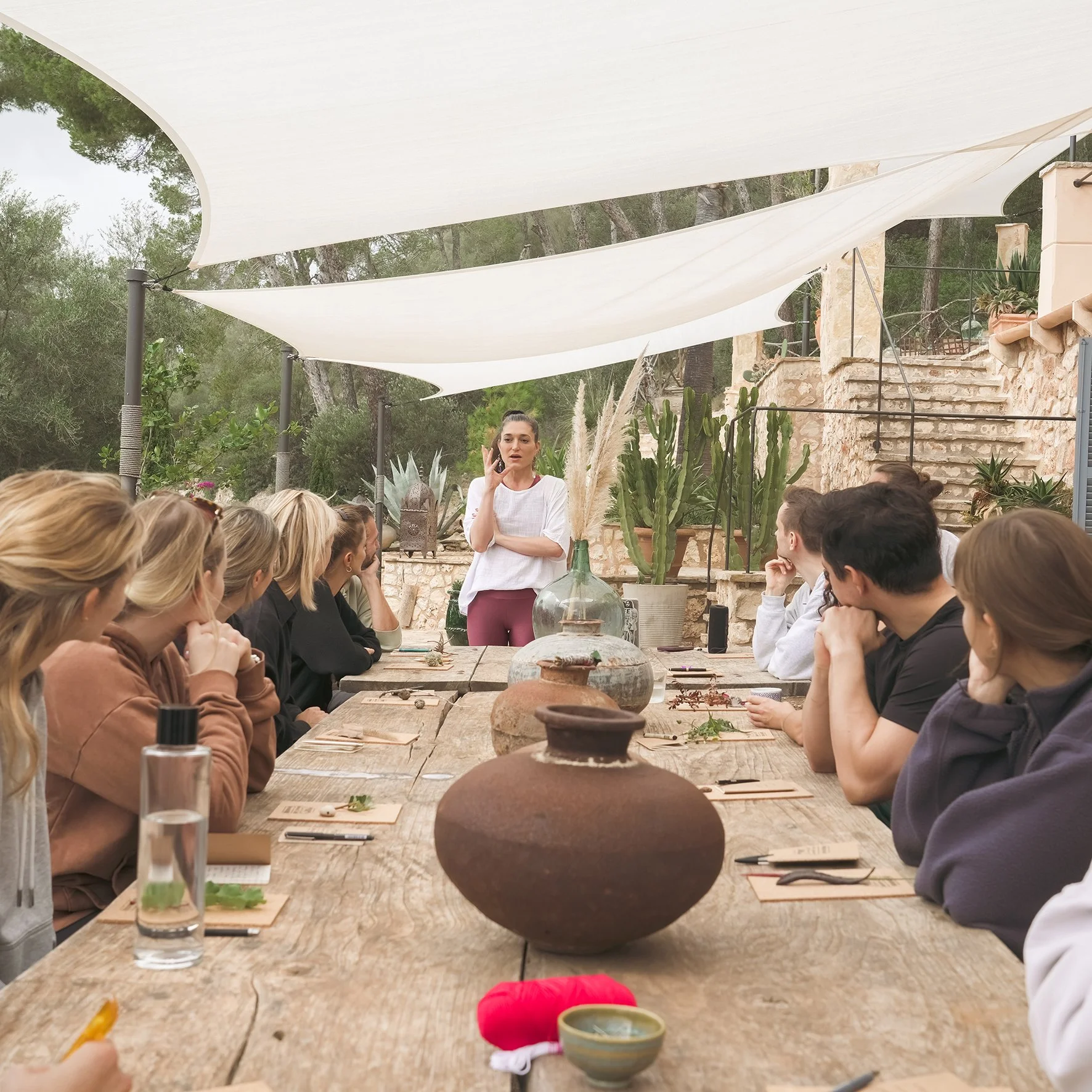 Menschen sitzen an einem langen Holztisch im Freien, während eine Frau vorne spricht. Es gibt Pflanzen im Hintergrund, darunter große Kakteen, und schattenspendende Segel über dem Tisch.