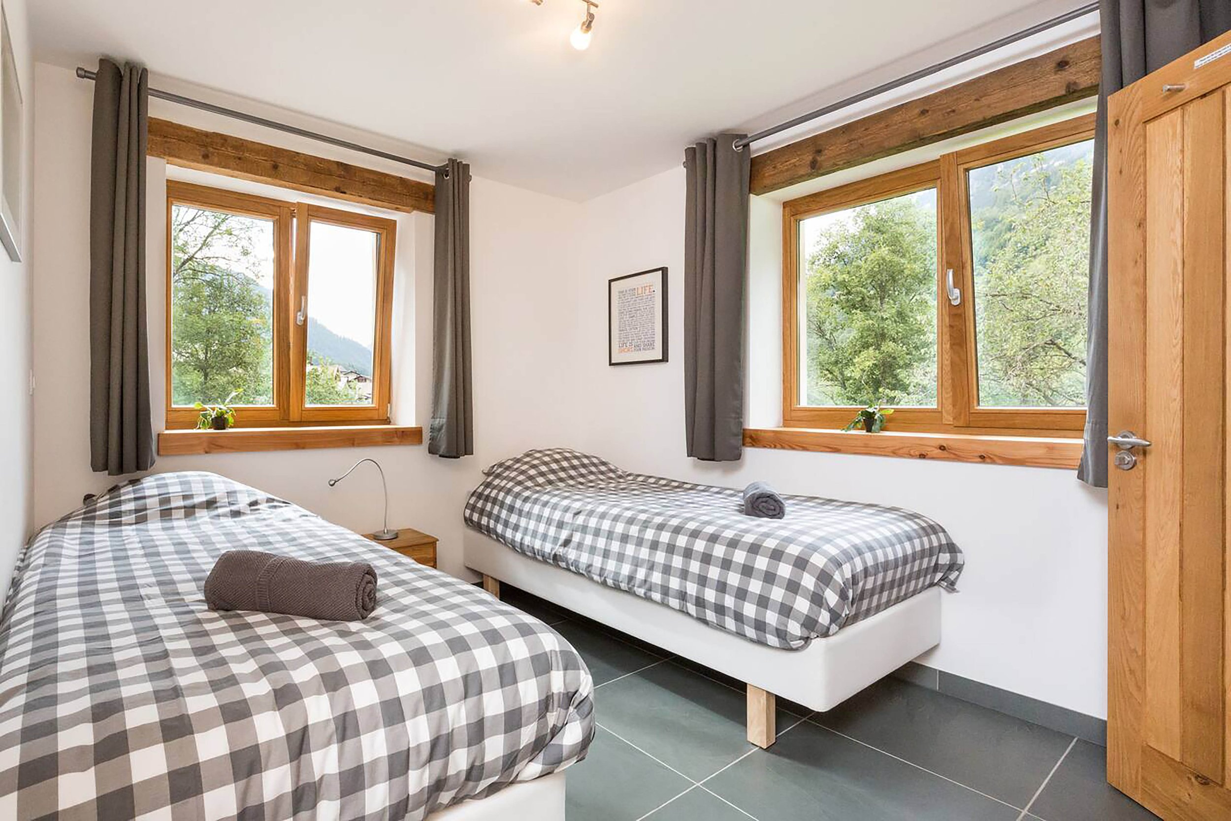 Ein helles Schlafzimmer mit zwei großen Fenstern, beige Vorhängen, einem kleinen Holztisch, drei Stühlen, einem Spiegel an der Wand und einem Bett.