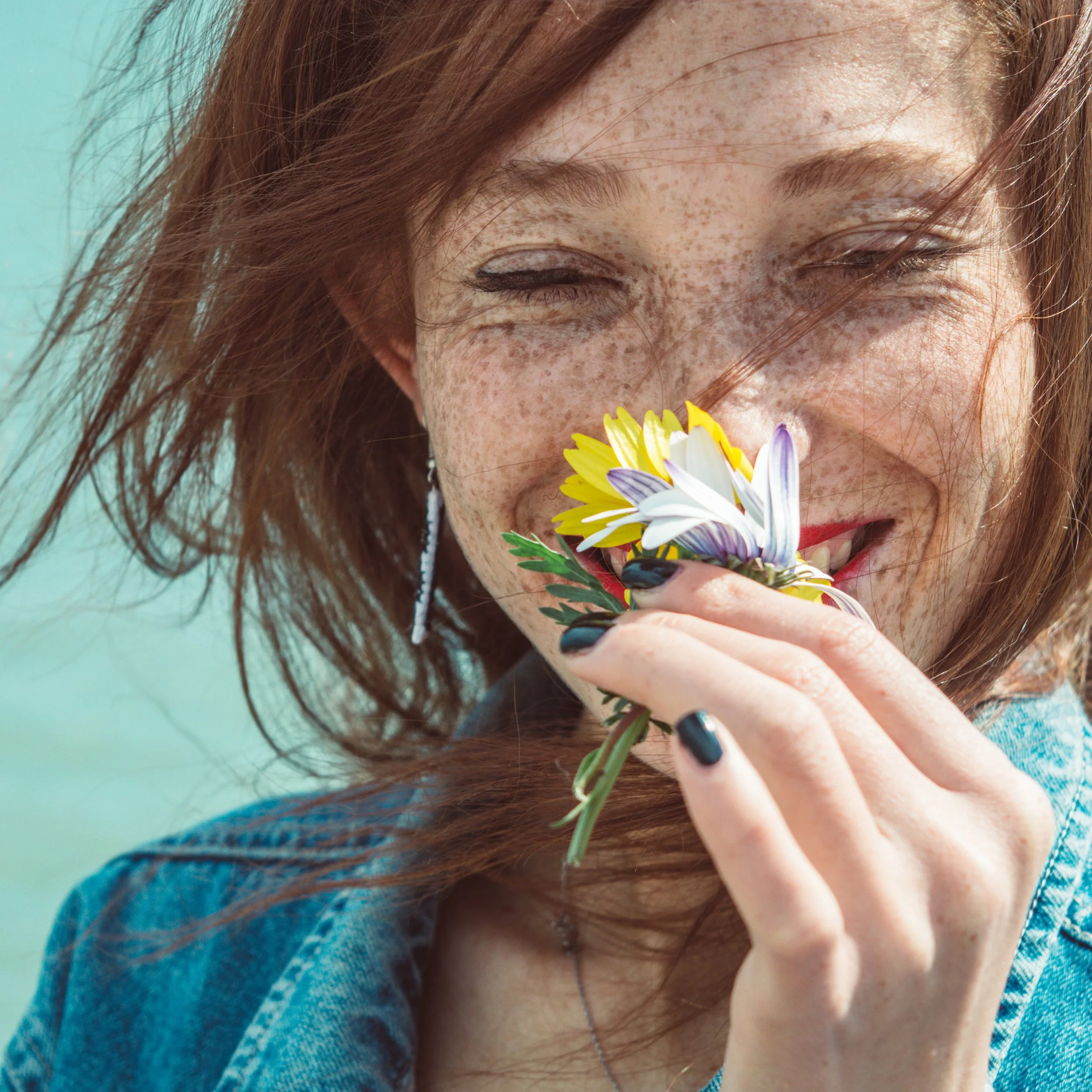 Lächelnde Frau mit Sommersprossen, die eine bunte Blumensträuße hält, mit geschlossen Augen, im Freien, bei Sonnenschein