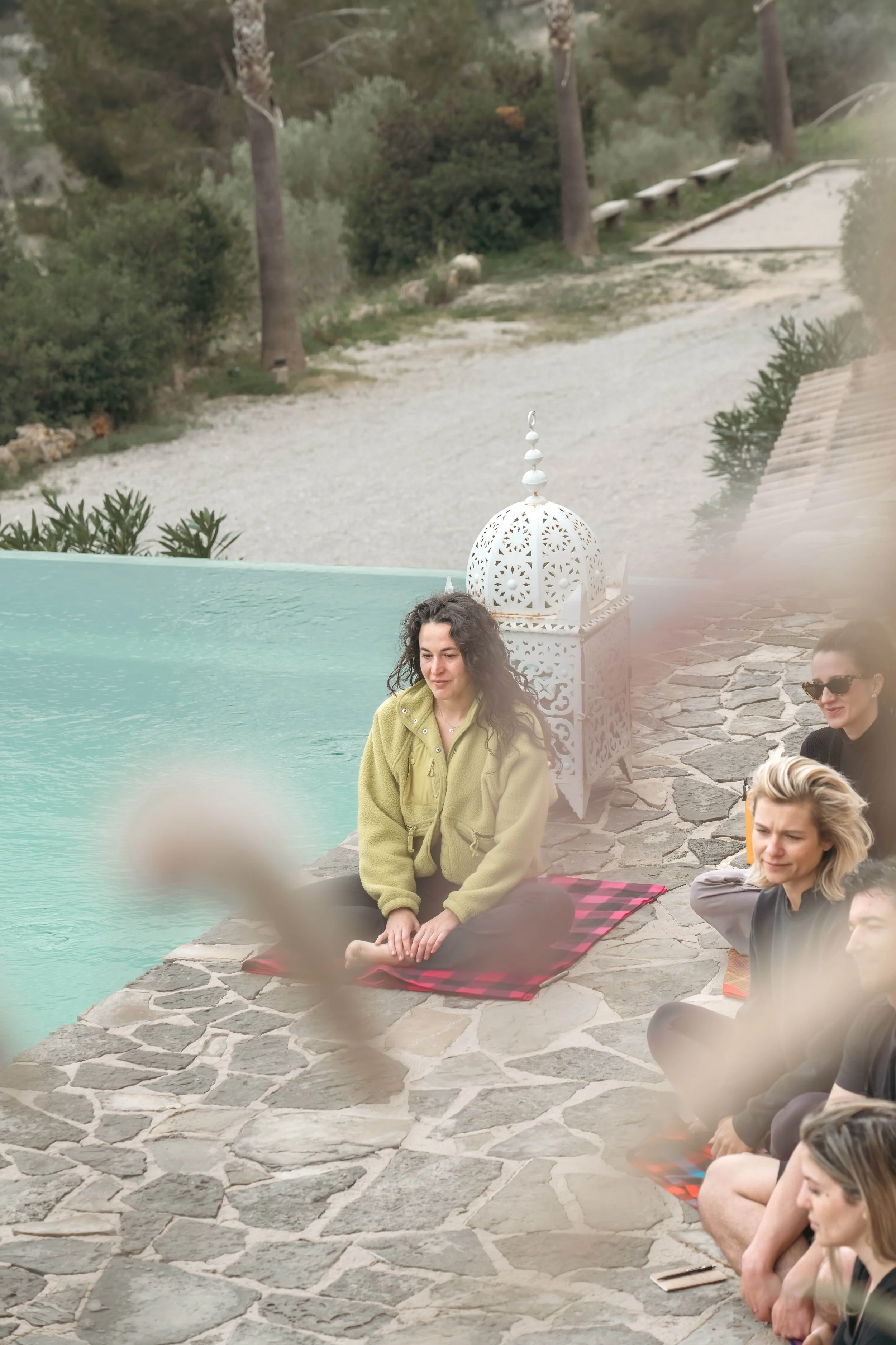Gruppe von Menschen, die am Pool im Freien sitzen, einige mit geschlossenen Augen, auf Tüchern auf dem Steinboden. Im Hintergrund sind Bäume und eine sandige Fläche sichtbar, die Gartenanlage hat eine orientalisch inspirierte laterneartige Dekoration.
