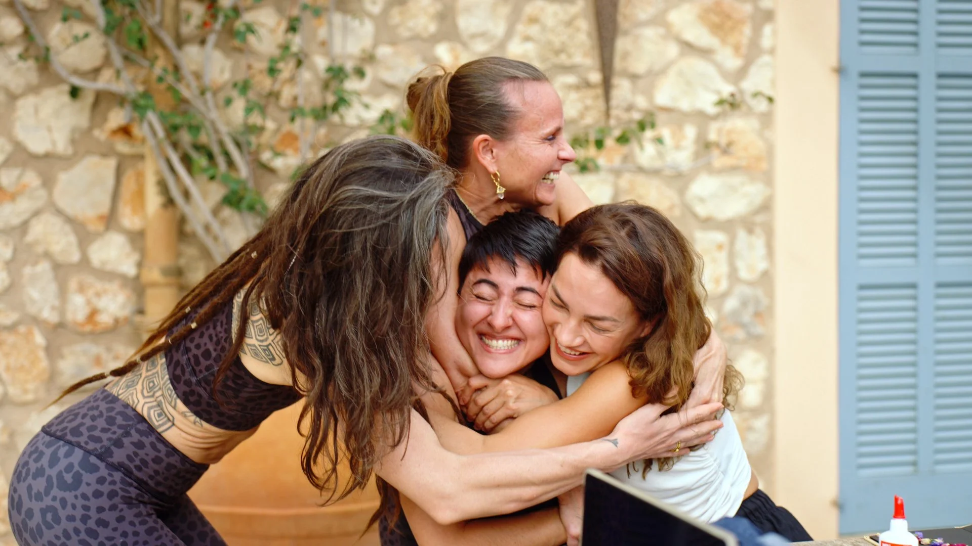 Vier Frauen lachen und umarmen sich freudig in einem Raum mit Steinwand und blauen Fensterläden.