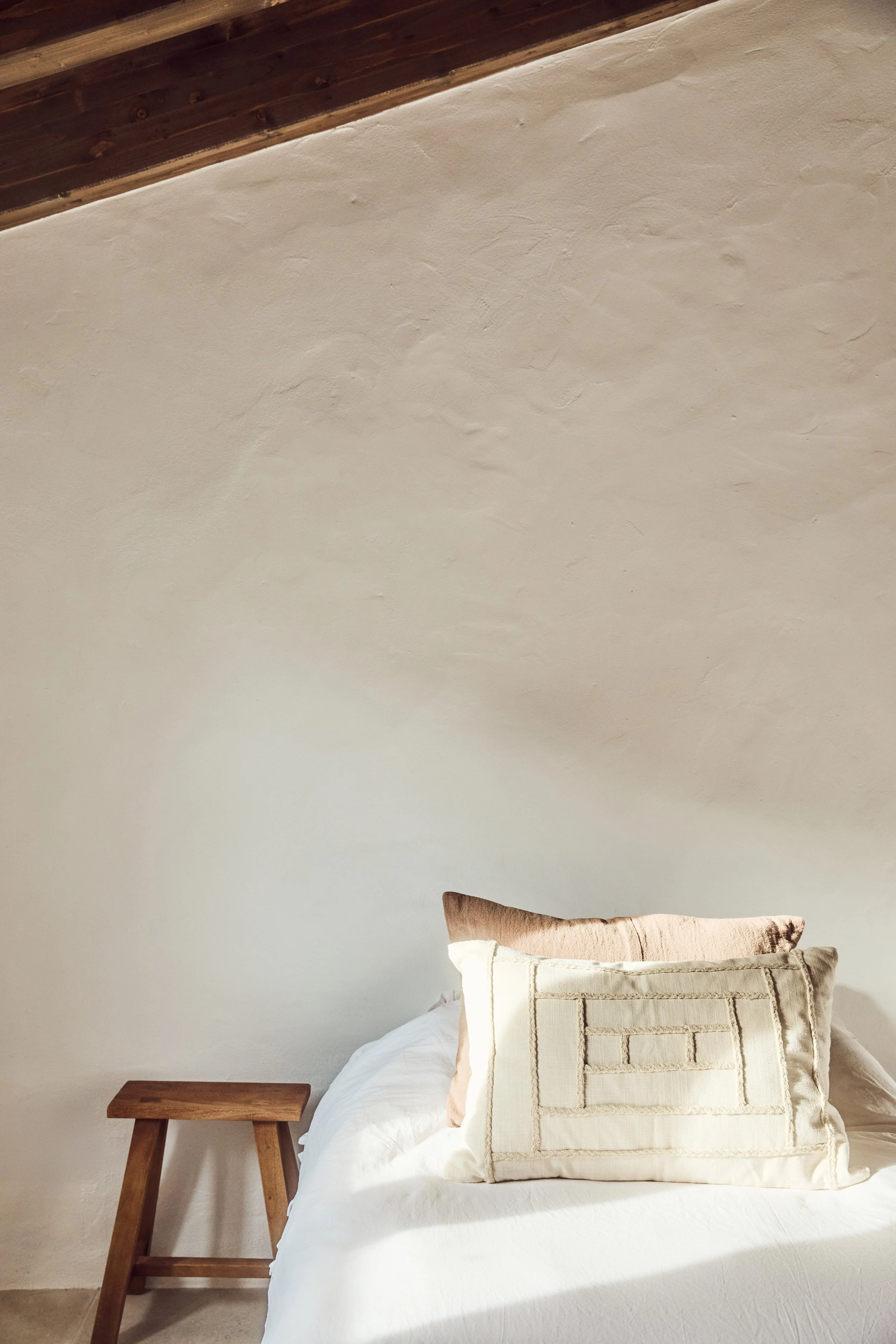 Ein Bett mit weißen Bettwäsche und zwei Kissen, eines in einem Beige-Ton und eines in einem Hellbraun-Ton, neben einem kleinen Holztisch in einem Raum mit einer weißen Wand und einer Holzdecke.
