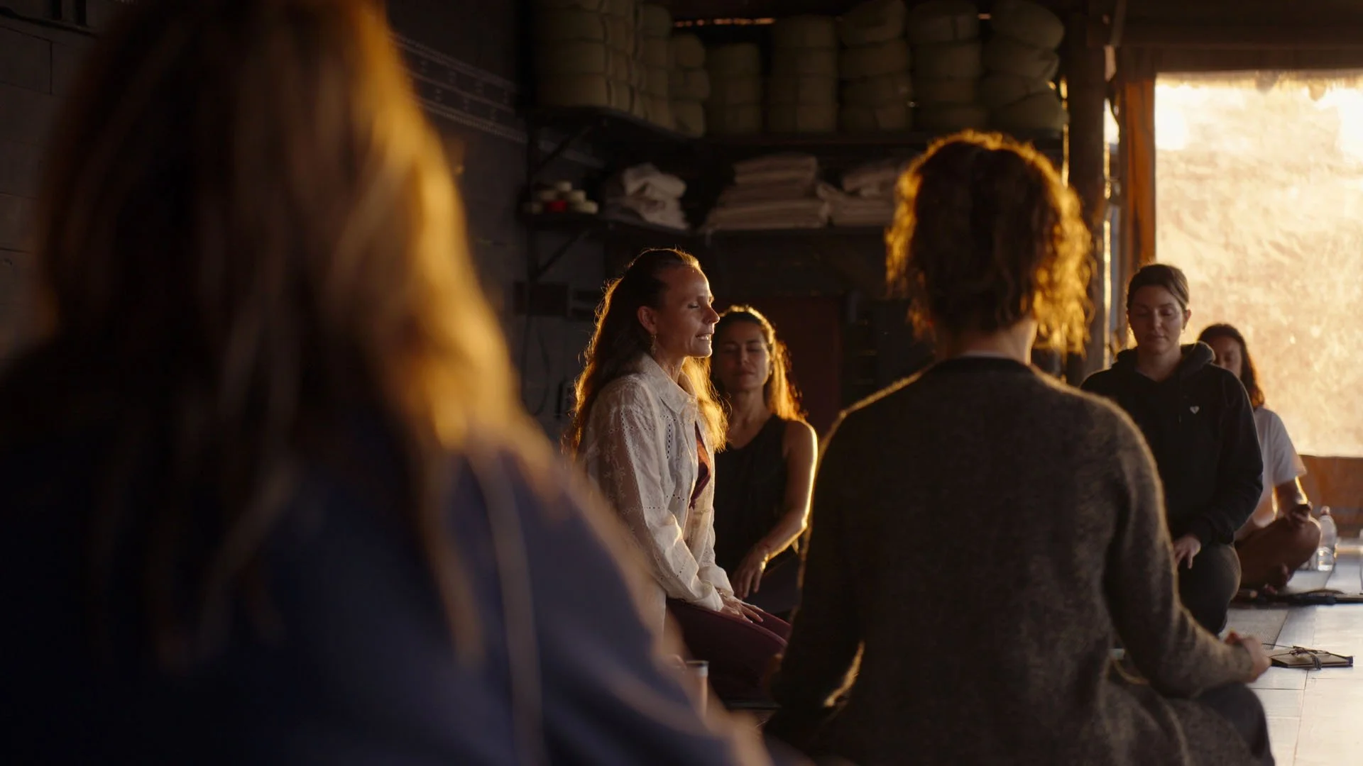 Menschen sitzen in einem Kreis in einem dunklen Raum, das Sonnenlicht scheint durch ein offenes Fenster im Hintergrund.