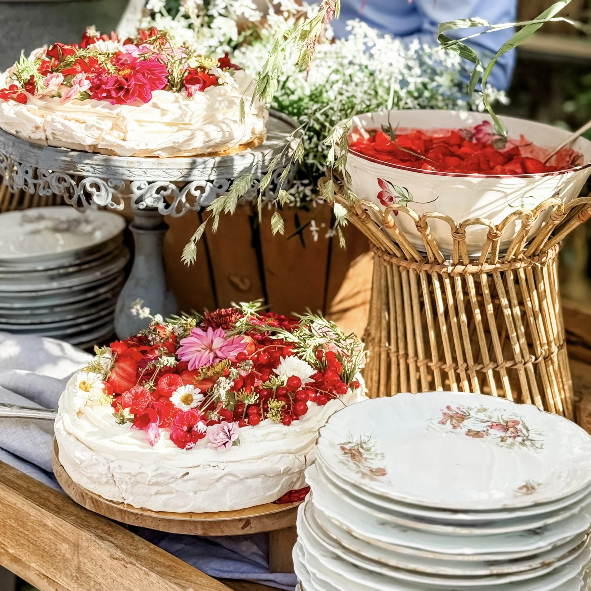 Oberteil eines gedeckten Tisches mit einem Obstkuchen, der mit roten und weißen Blumen und Beeren dekoriert ist, einer Schüssel mit roten Früchten in einem Korb, einem Blumenmustergeschirrset und einem Stapel weißer Teller.