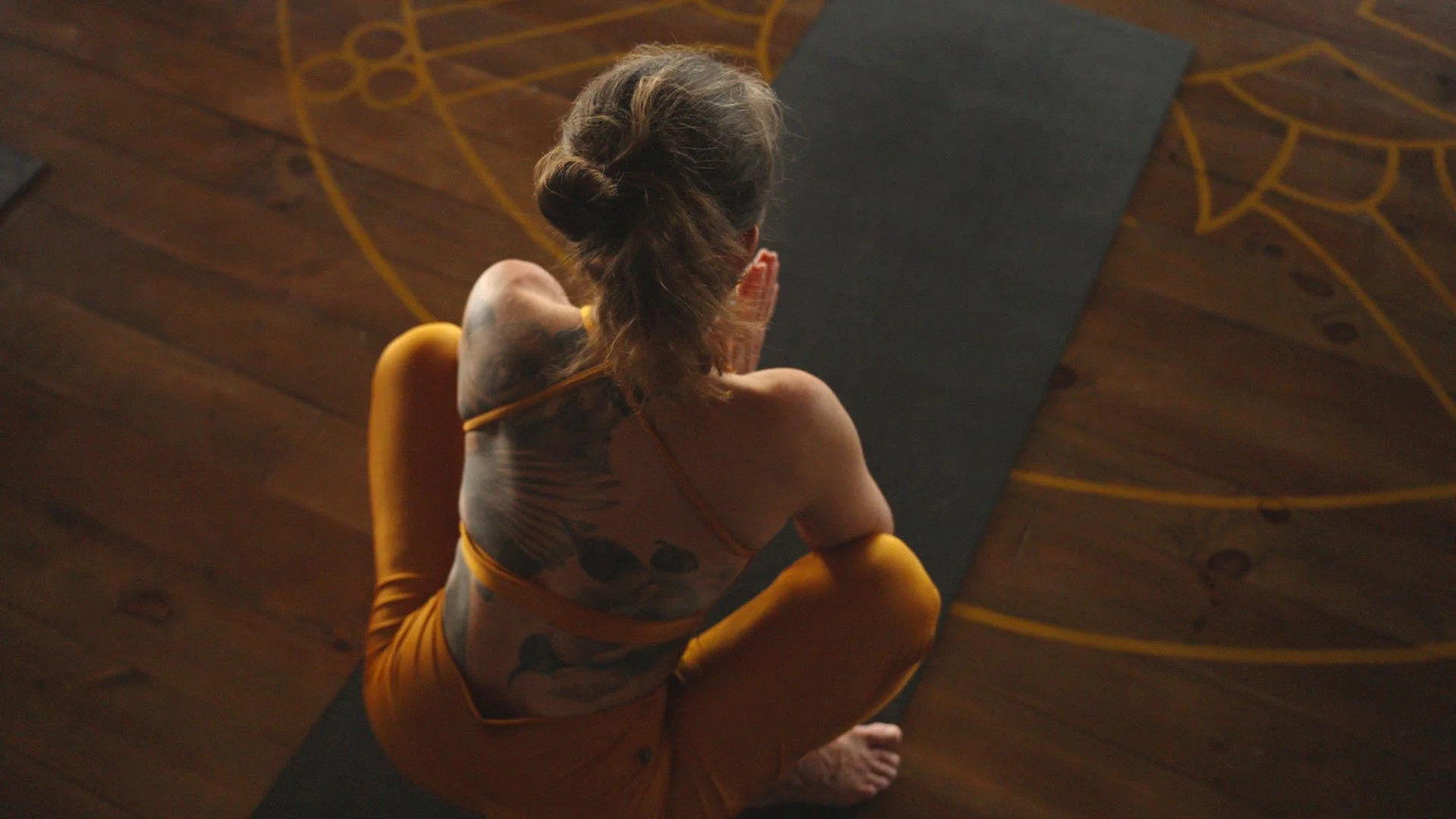 Frau beim Yoga auf einem schwarzen Yogaboden, umgeben von Holzparkett mit goldfarbenen Yoga-Symbolen.