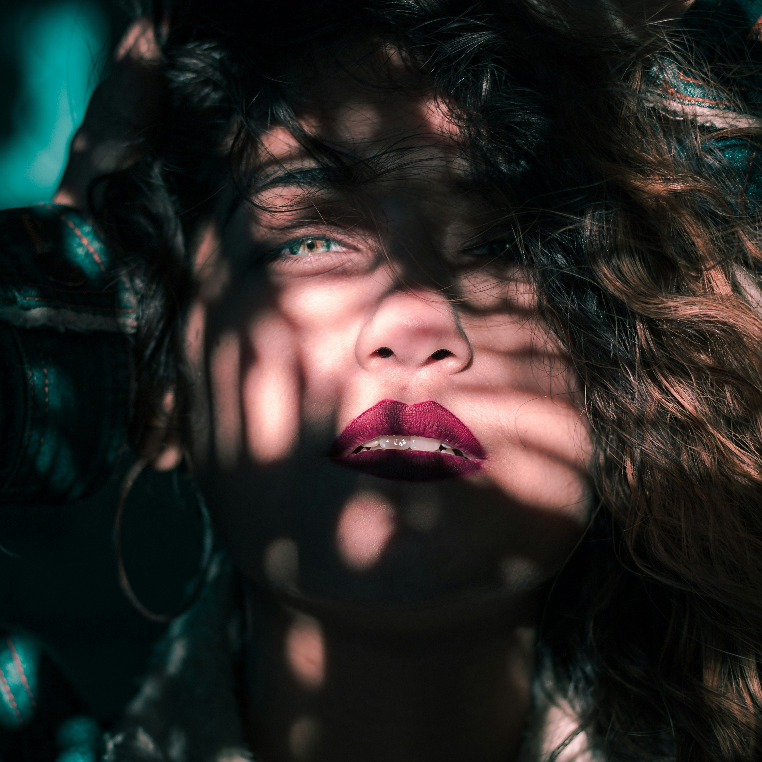 Nahaufnahme eines Gesichts mit lockigem Haar, das durch Schatten von Blättern aufgeteilt ist, mit betonten roten Lippen und grünen Augen.