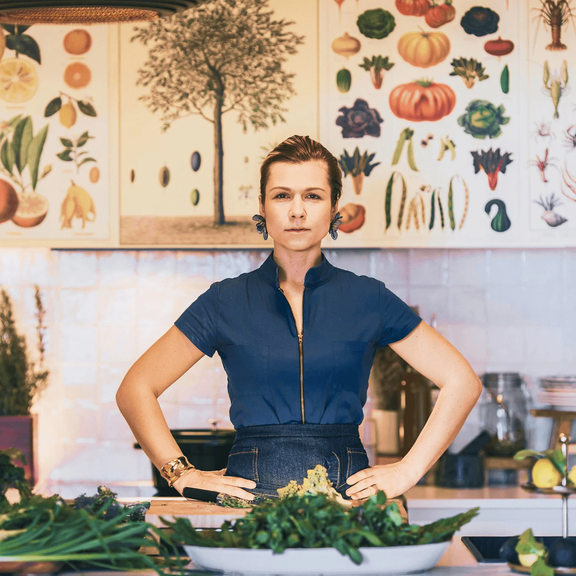 Eine Frau steht in einer Küche mit starkem Blick in die Kamera, umgeben von frischem Gemüse. Hinter ihr sind Gemälde mit Gemüse und Pflanzen an der Wand.