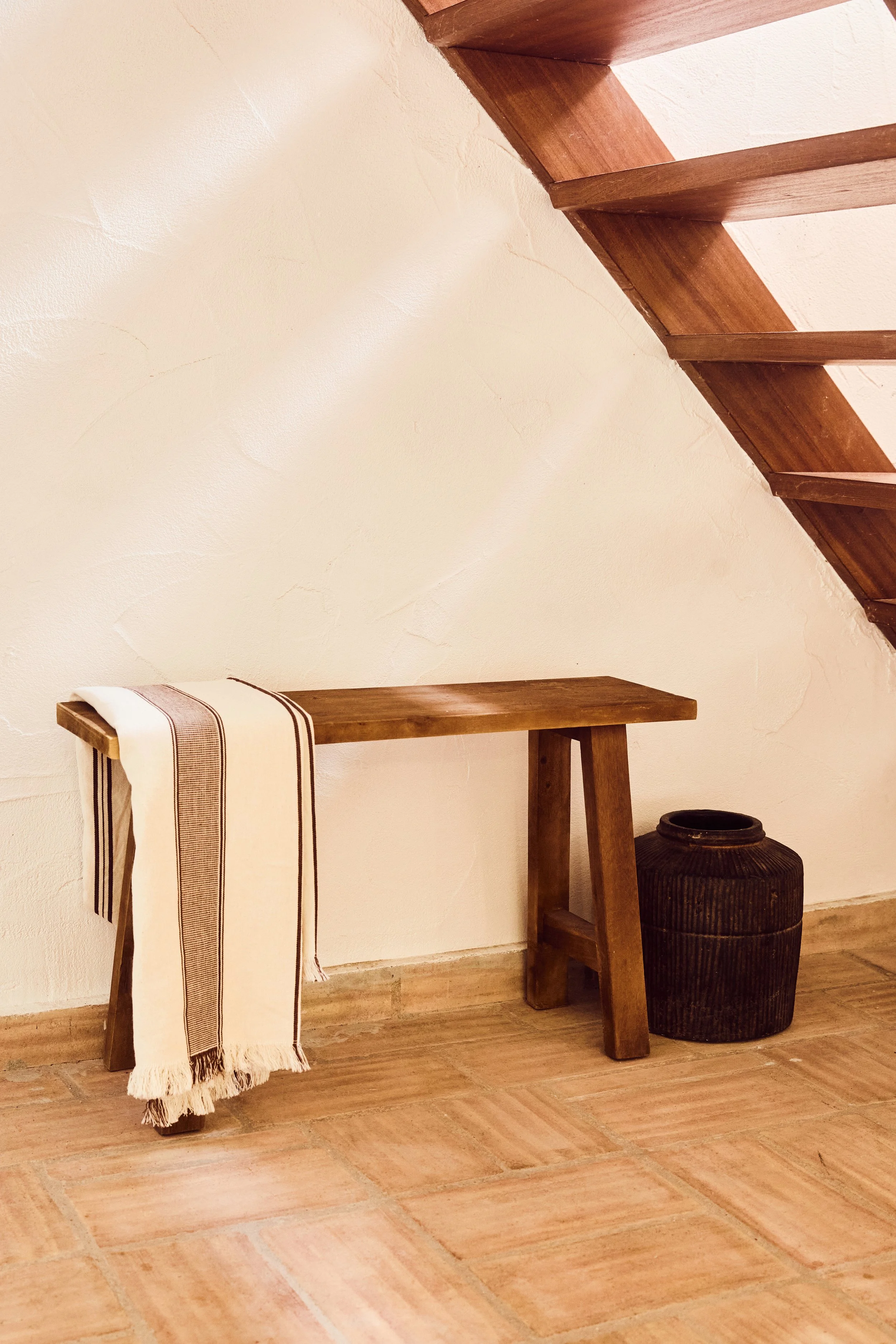 Ein Holzbank mit einem weißen und braunen gestreiften Tuch hängt an der linken Seite. Daneben steht eine runde, dunkle, geflochtene Vase auf dem Boden. Im Hintergrund links ist eine weiße Wand, an der eine Holztreppe seitlich nach oben führt.
