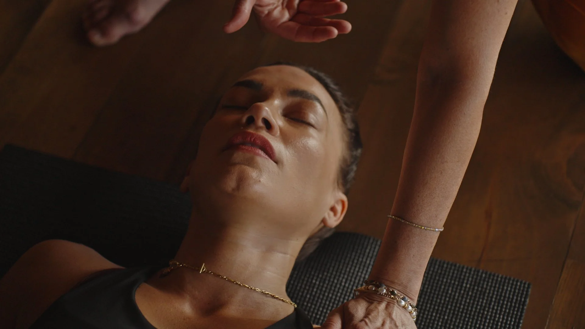 Eine Frau liegt entspannt auf dem Rücken auf einer Matte, während eine andere Person ihr eine Massage gibt. Die Frau hat geschlossene Augen, trägt eine schwarze Kleidung und Schmuck, und hat dunkle, glatte Haare.