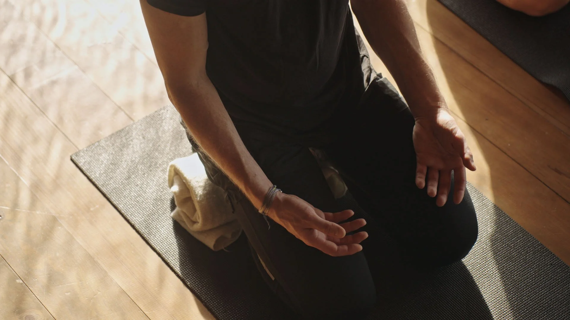 Eine Person, die im Schneidersitz auf einer Yogamatte im Schatten sitzt, die Hände offen auf den Oberschenkeln. Es ist eine helle Umgebung mit Holzboden, und die Person trägt ein dunkles T-Shirt.