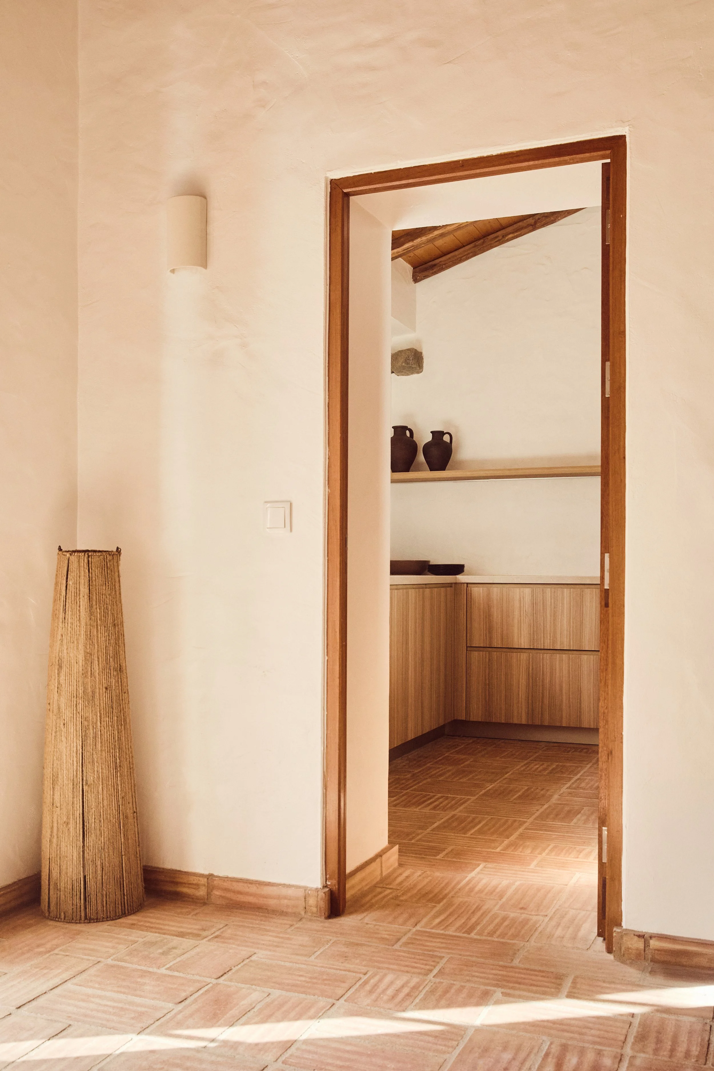 Ein Blick durch eine offene Tür in eine Küche mit Holzverkleidung, dunklen Töpfen auf einem Regal und einem Gegenstand in einer Ecke.