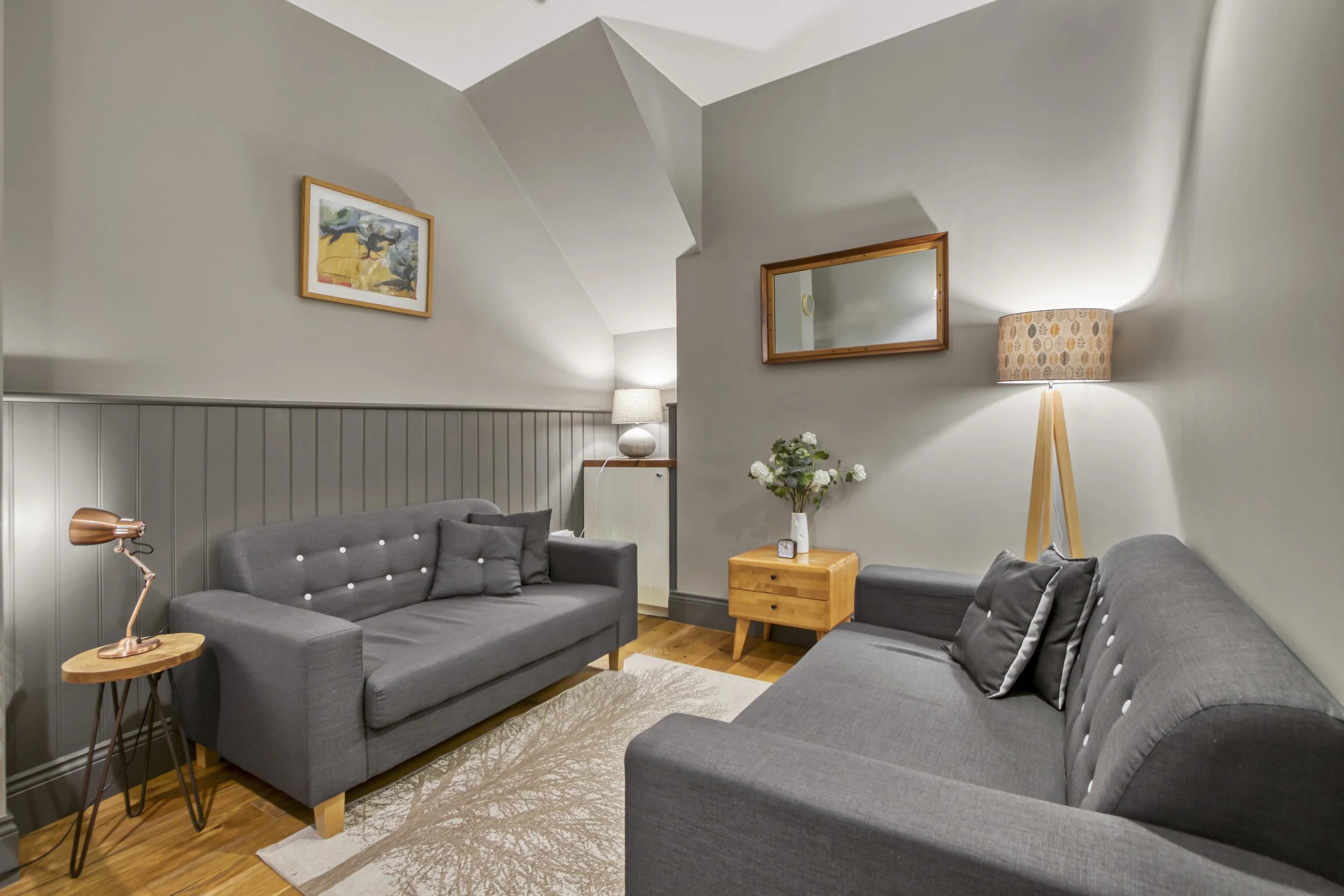 Living room with two gray sofas, a wooden side table, a table lamp, a floor lamp, wall art, a mirror, a vase of white flowers, and a beige rug.