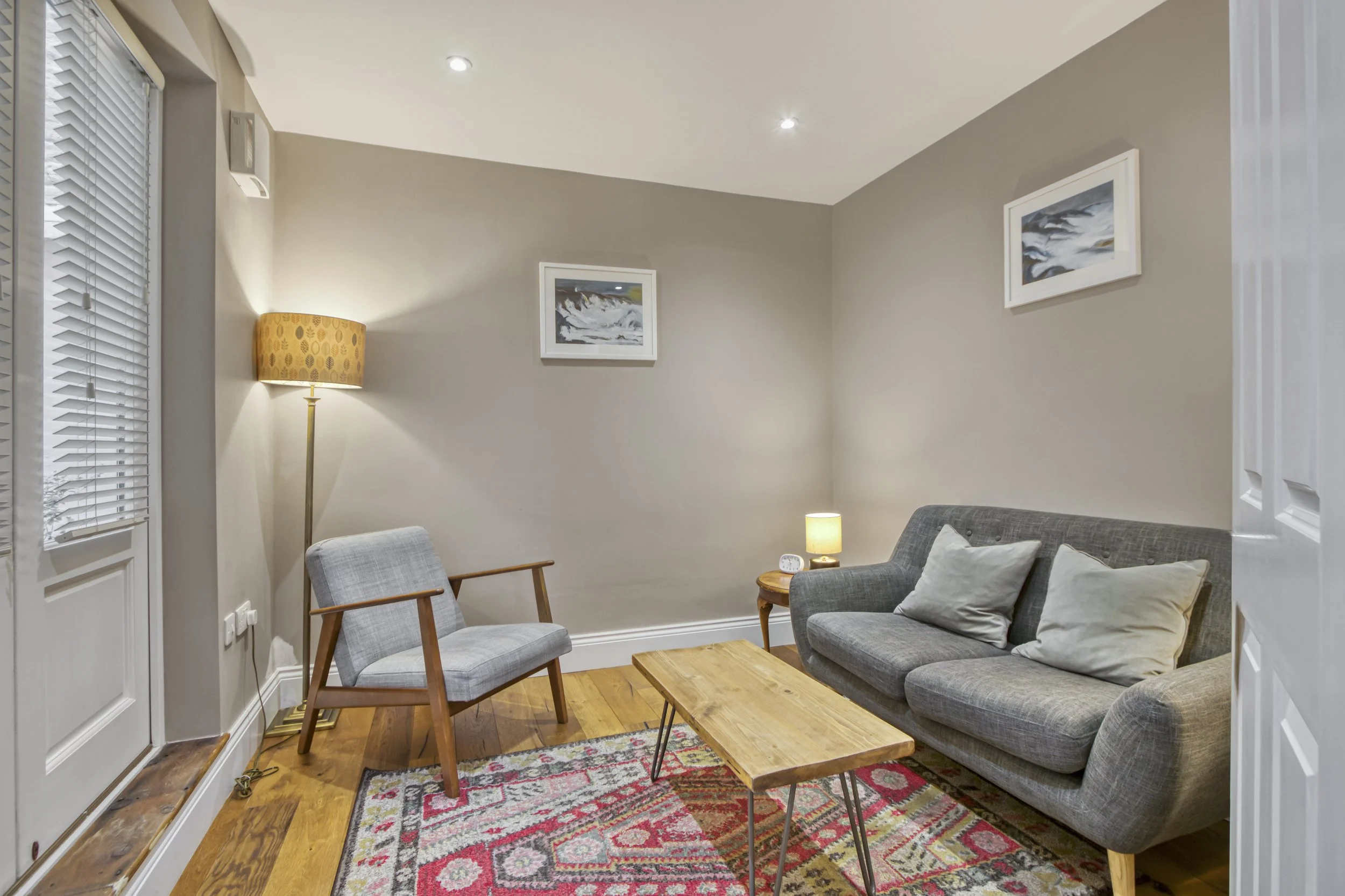 Living room with a gray sofa and cushions, a gray armchair, a wooden coffee table, a small wooden side table with a lamp, a tall floor lamp, framed artwork on the wall, and a colorful patterned rug on hardwood floor.