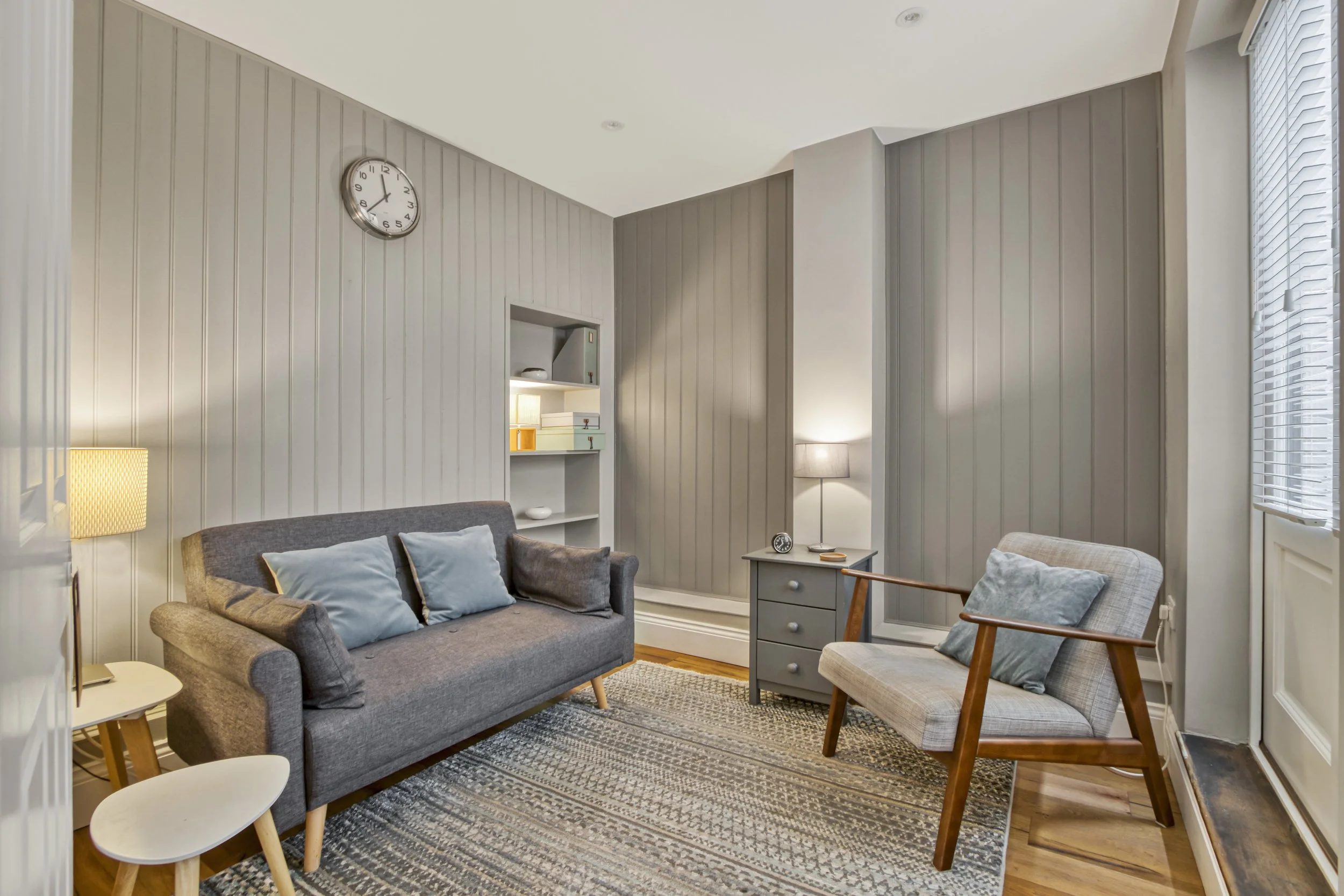 Cozy living room with gray sofa, wooden armchair with light-colored upholstery, side table, floor lamp, wall clock, and built-in shelves with decorative items. The room has gray paneled walls, wooden flooring, and large windows with blinds.