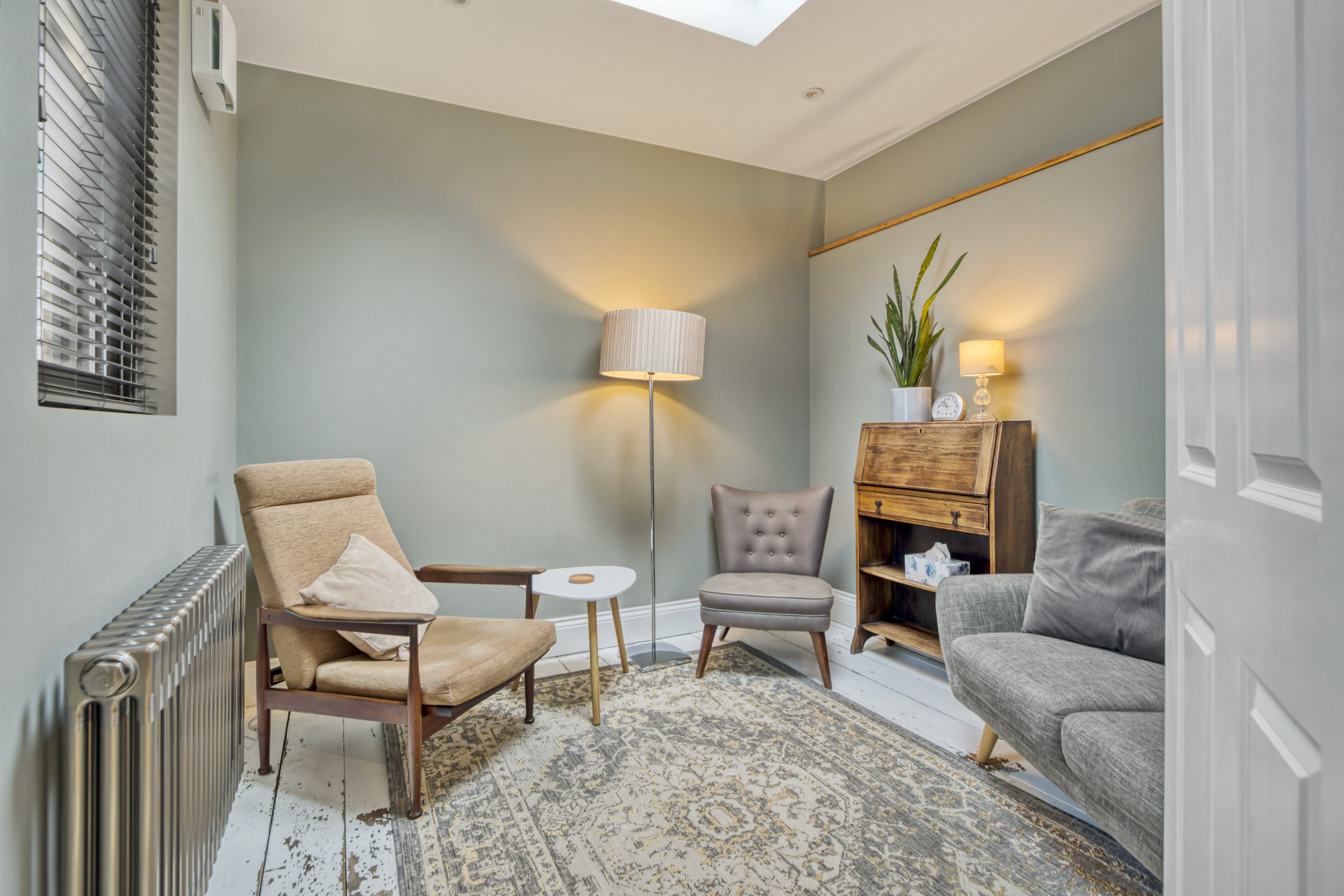 A cozy living room with grey and beige furniture, including a beige armchair, a grey armchair, and a grey sofa. The room features a patterned area rug, a tall floor lamp, a wooden cabinet with a plant and a small lamp on top, and a window with blinds.