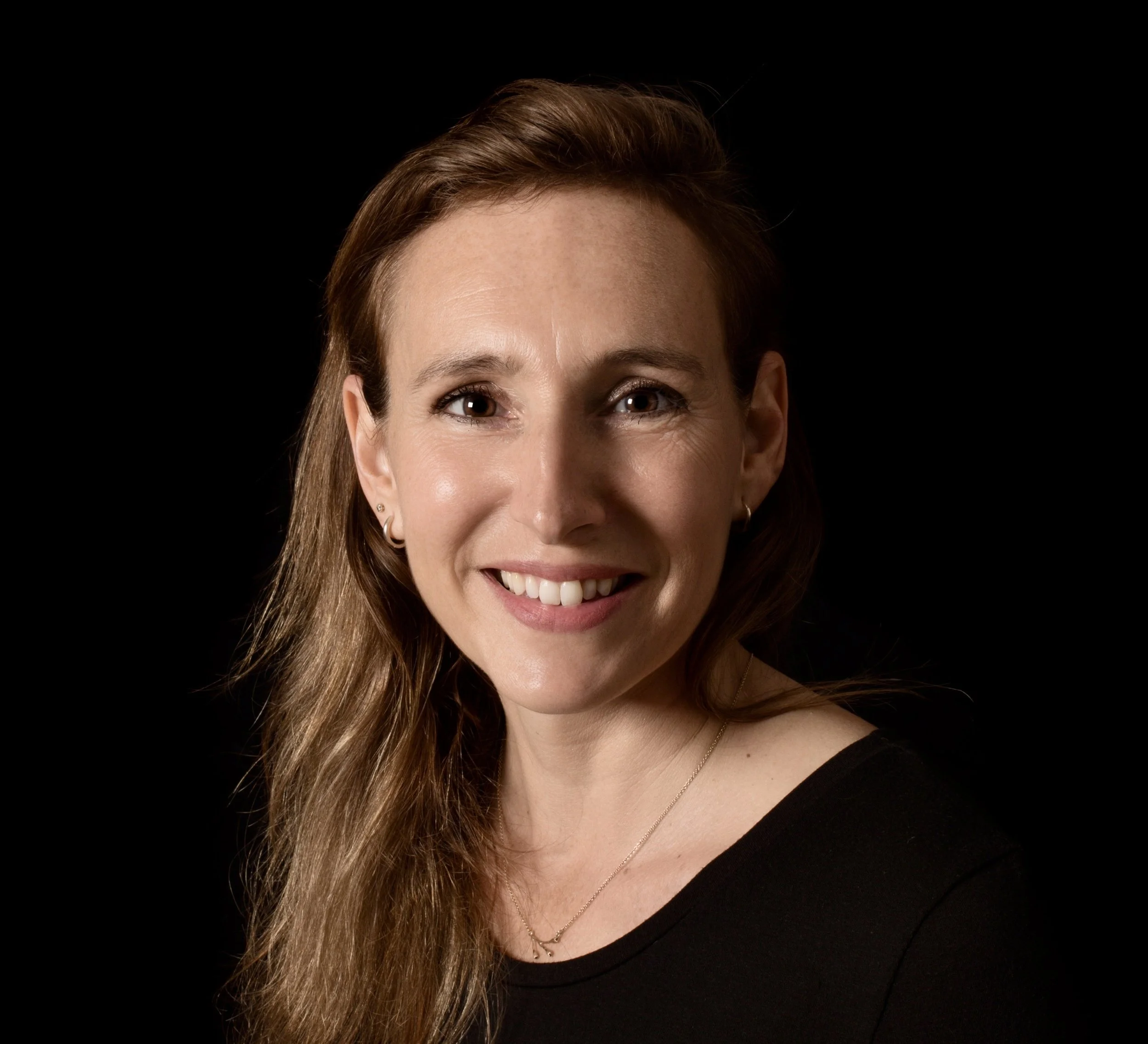 Smiling woman with long wavy light brown hair, wearing a black top and small earrings, against a black background.