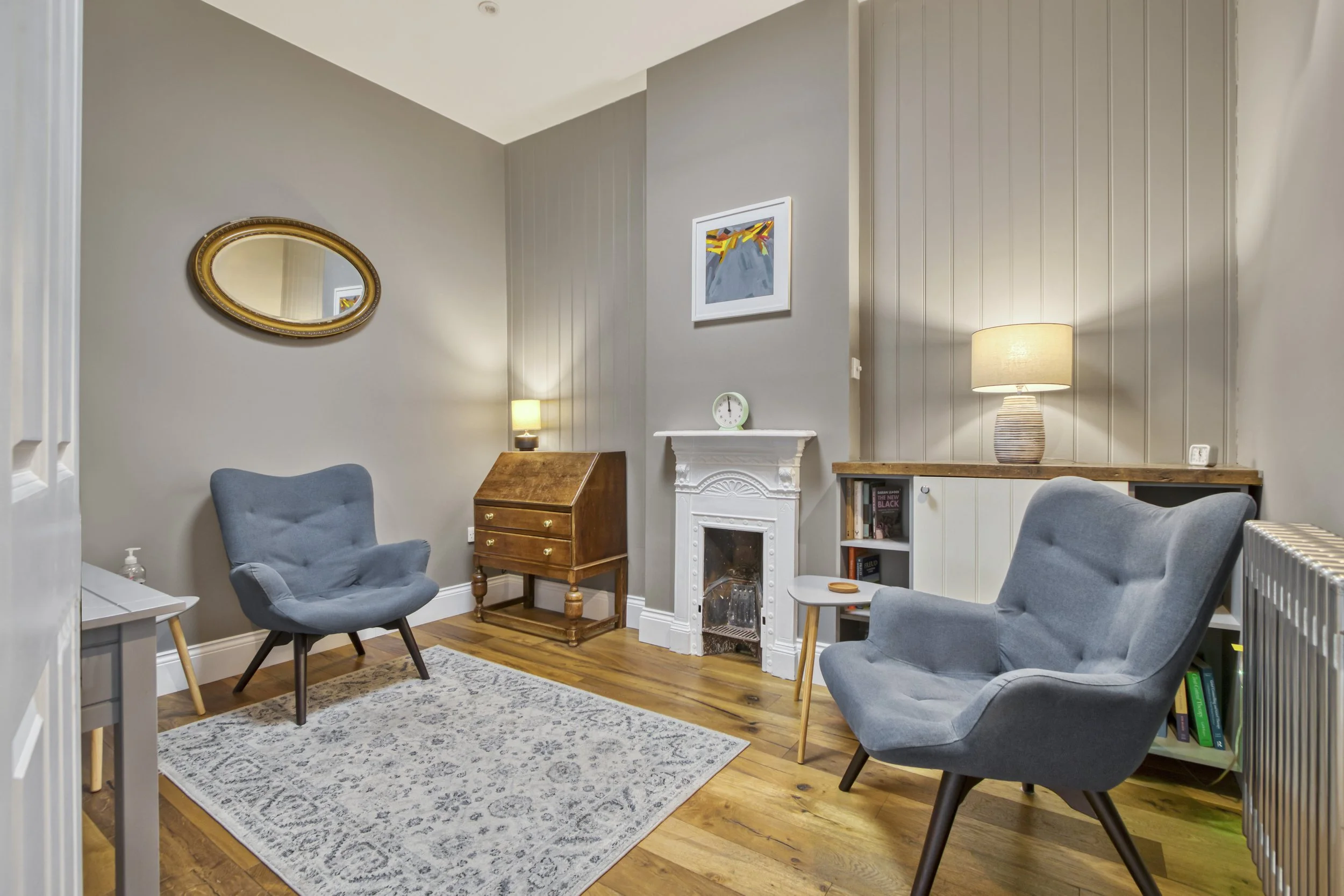 Living room with two gray chairs, small white side table, wooden cabinet with lamp, fireplace, framed artwork, mirror, bookshelf, and lamps on the walls.