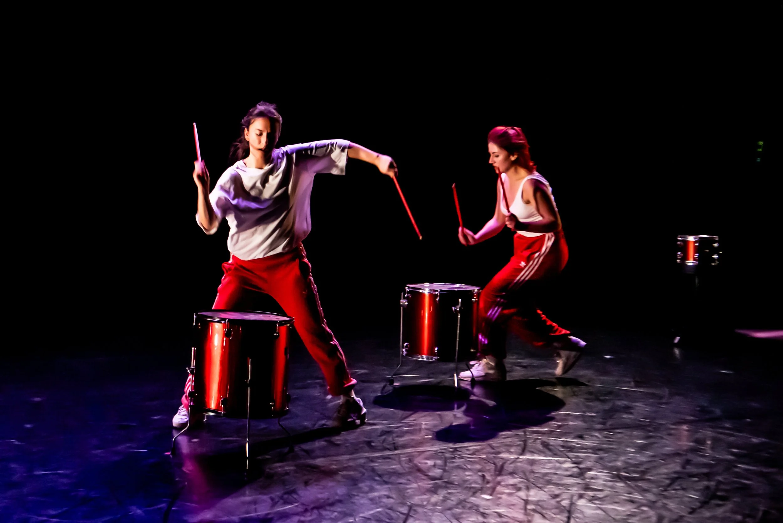Two performers dressed in white tops and red trousers are captured in energetic motion, one front facing, the other in profile, holding red drumsticks about to hit free standing drums, a drum kit dispersed within the black box space.