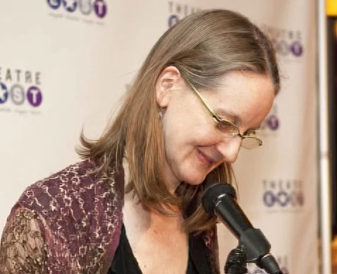A person speaking into a microphone at a theatre event with a backdrop featuring a logo and text.
