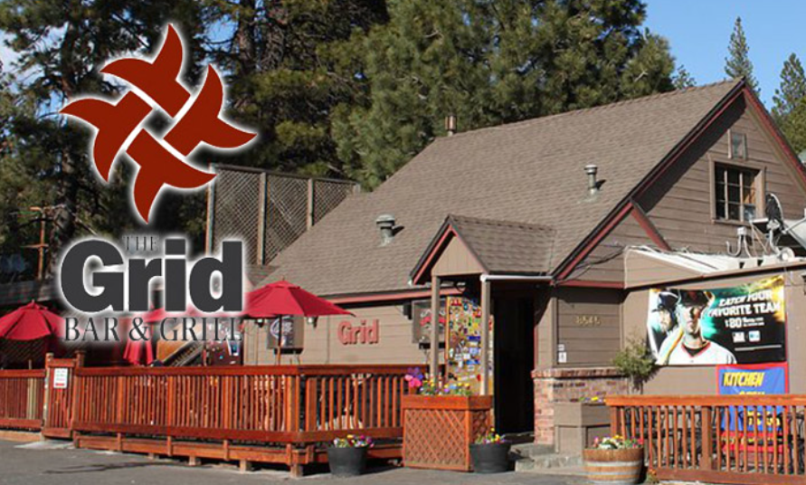 Exterior of The Grid Bar & Grill with outdoor seating, red umbrellas, and a sign featuring a baseball player