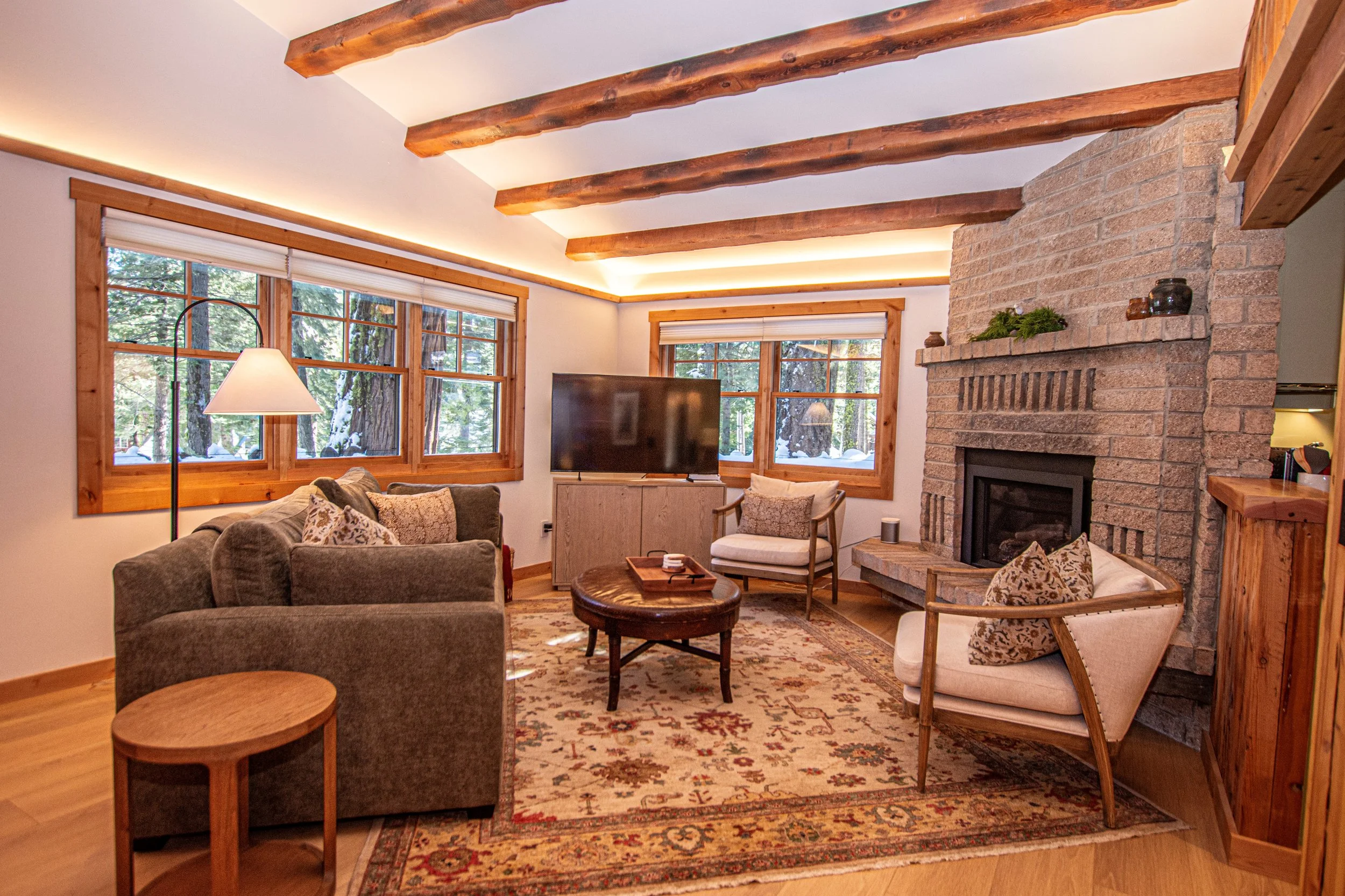 Living room with a fireplace, large windows, sofa, armchair, wooden coffee table, rug, and a television, surrounded by wooden beams and trim.
