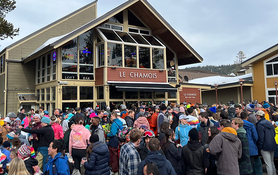 Crowd of people outside Le Chamois restaurant in a ski resort village, with mountains and ski slopes in the background.