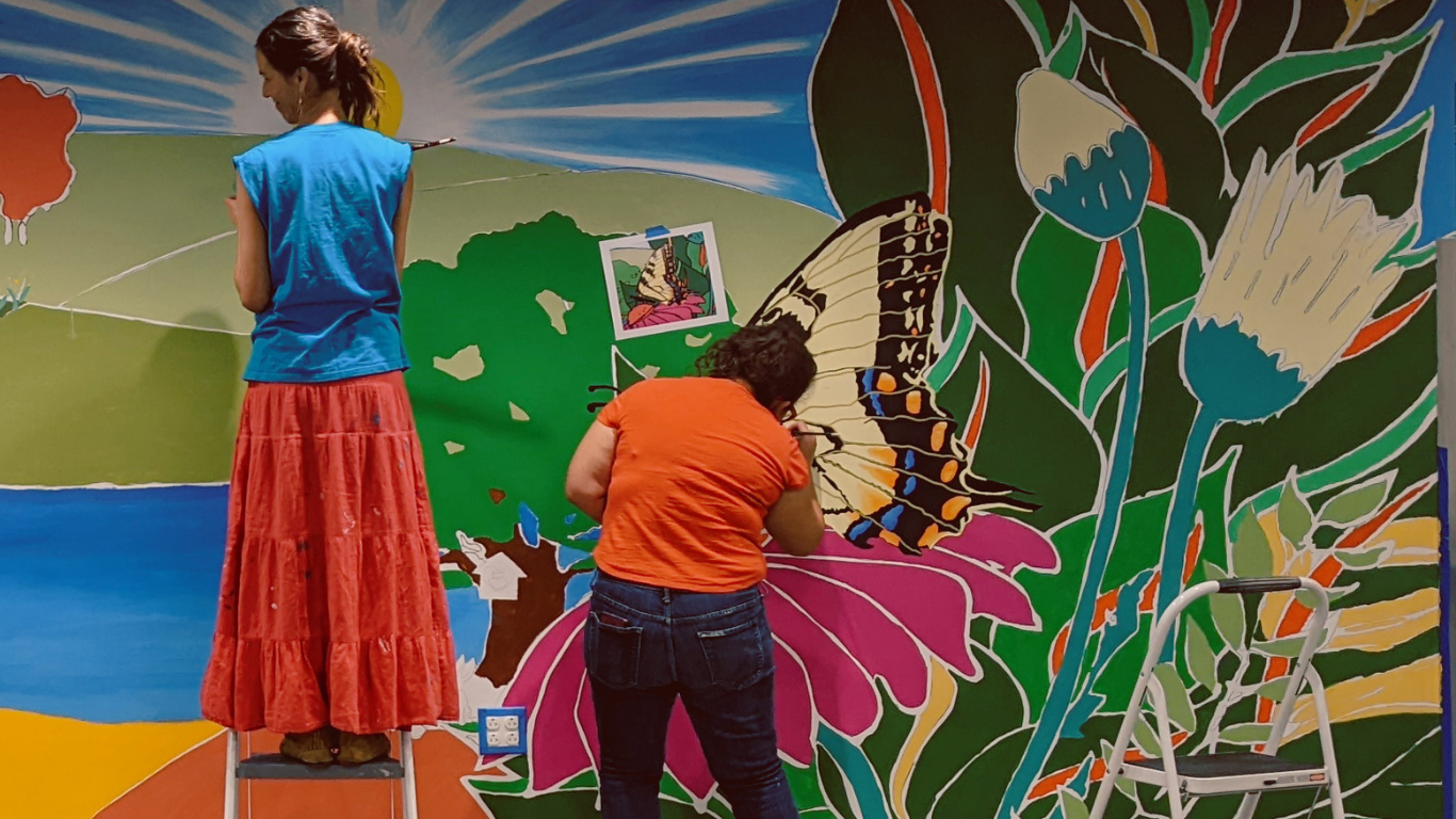 Volunteers for an Art Room Community Art Project donate a mural to fellow Denton nonprofit Our Daily Bread. Two people stand in front of the colorful mural they are painting, which depicts flowers, hills, a tree, and a blue sky with sunbeams.