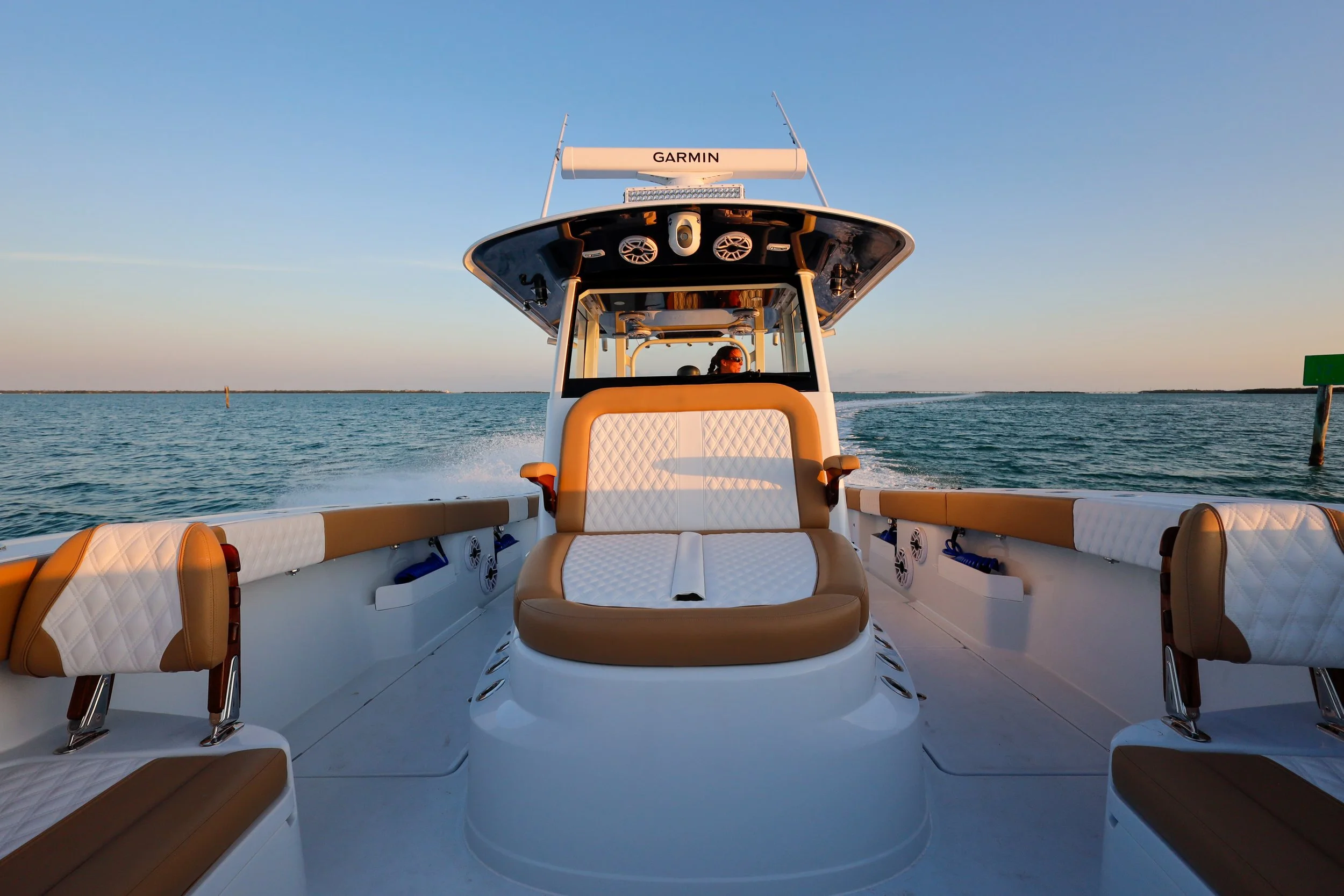 View from the front of a luxury motorboat on calm water during sunset, showing the boat's beige and white cushioned seating and the open sea in the background.