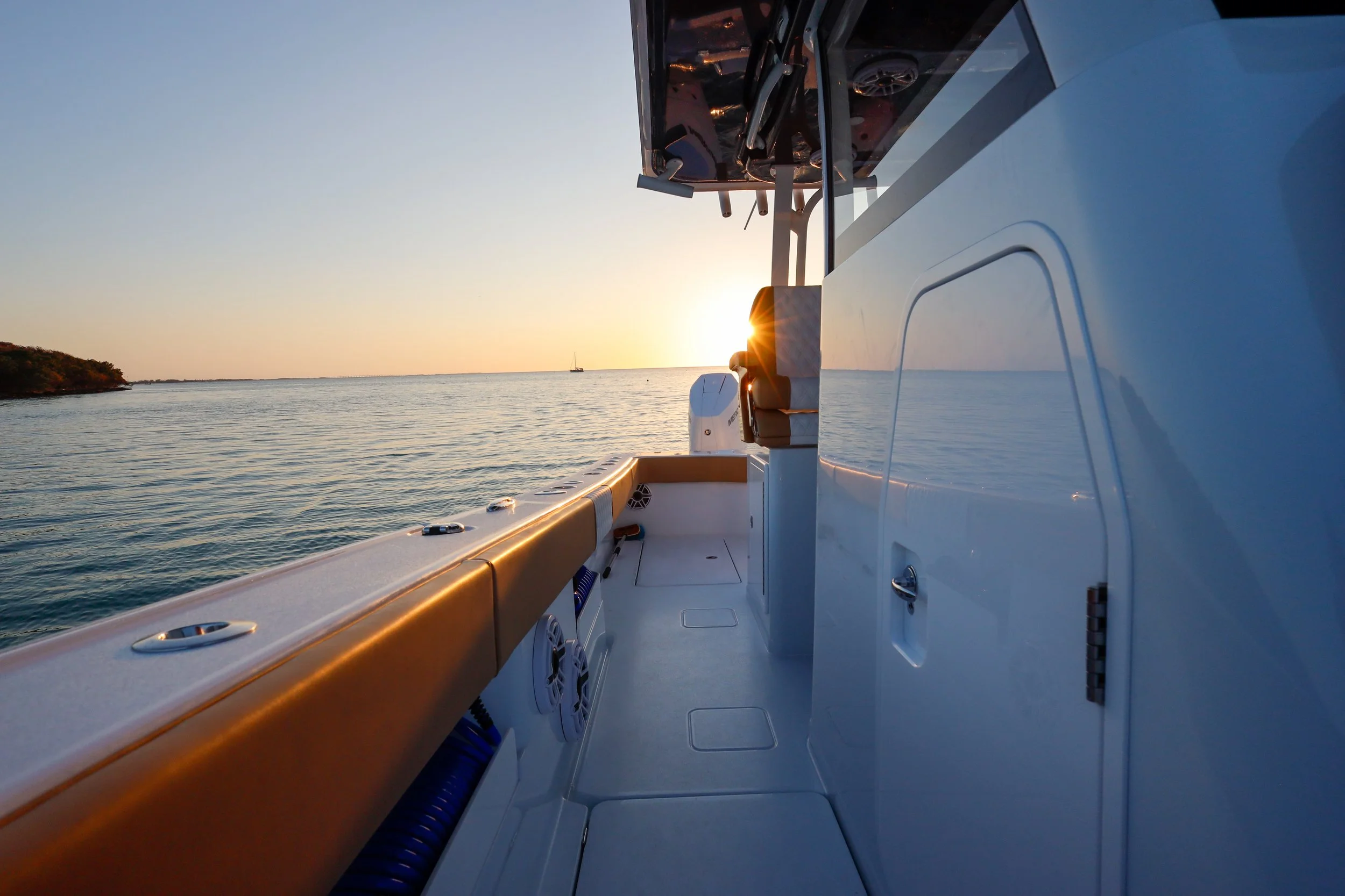 View from a boat at sunset showing a calm ocean, with a distant sailboat, and a shoreline with trees on the left.