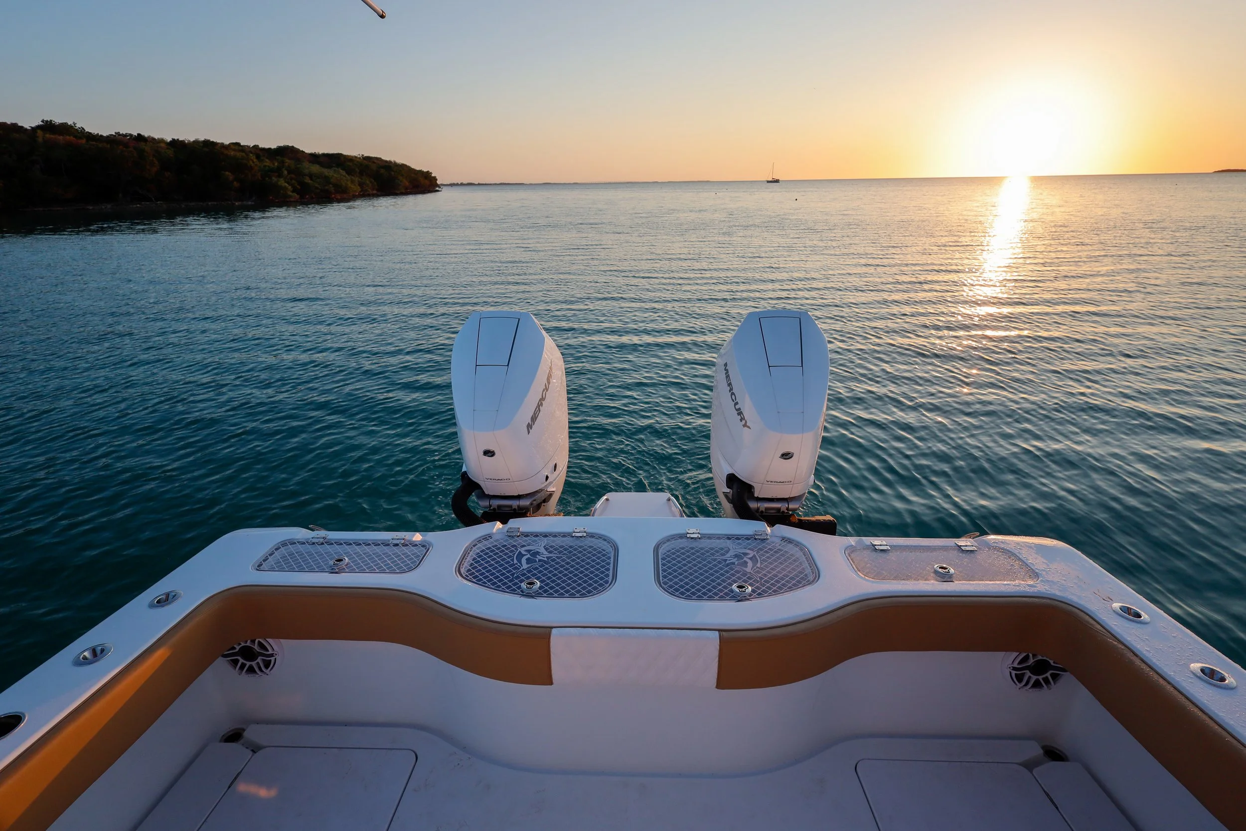 View from the back of a boat with two Mercury outboard motors, overlooking calm water at sunset with a distant landmass and a sailboat on the horizon.